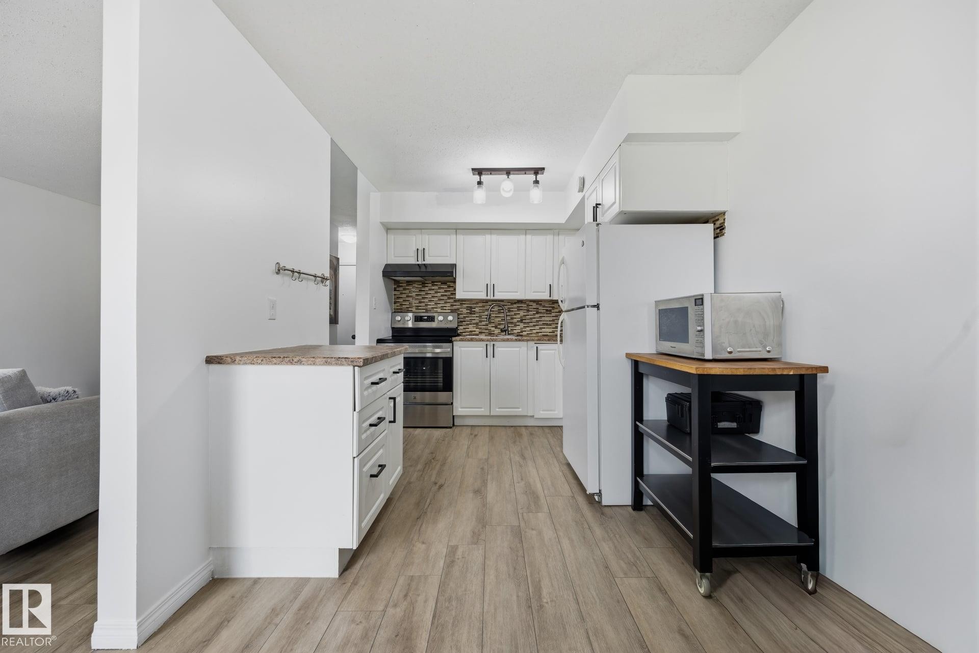Contemporary kitchen featuring white cabinetry, a stone-look backsplash, stainless steel appliances, wood-finish flooring, and a track lighting fixture - 105 16428 109 Street, Edmonton, AB