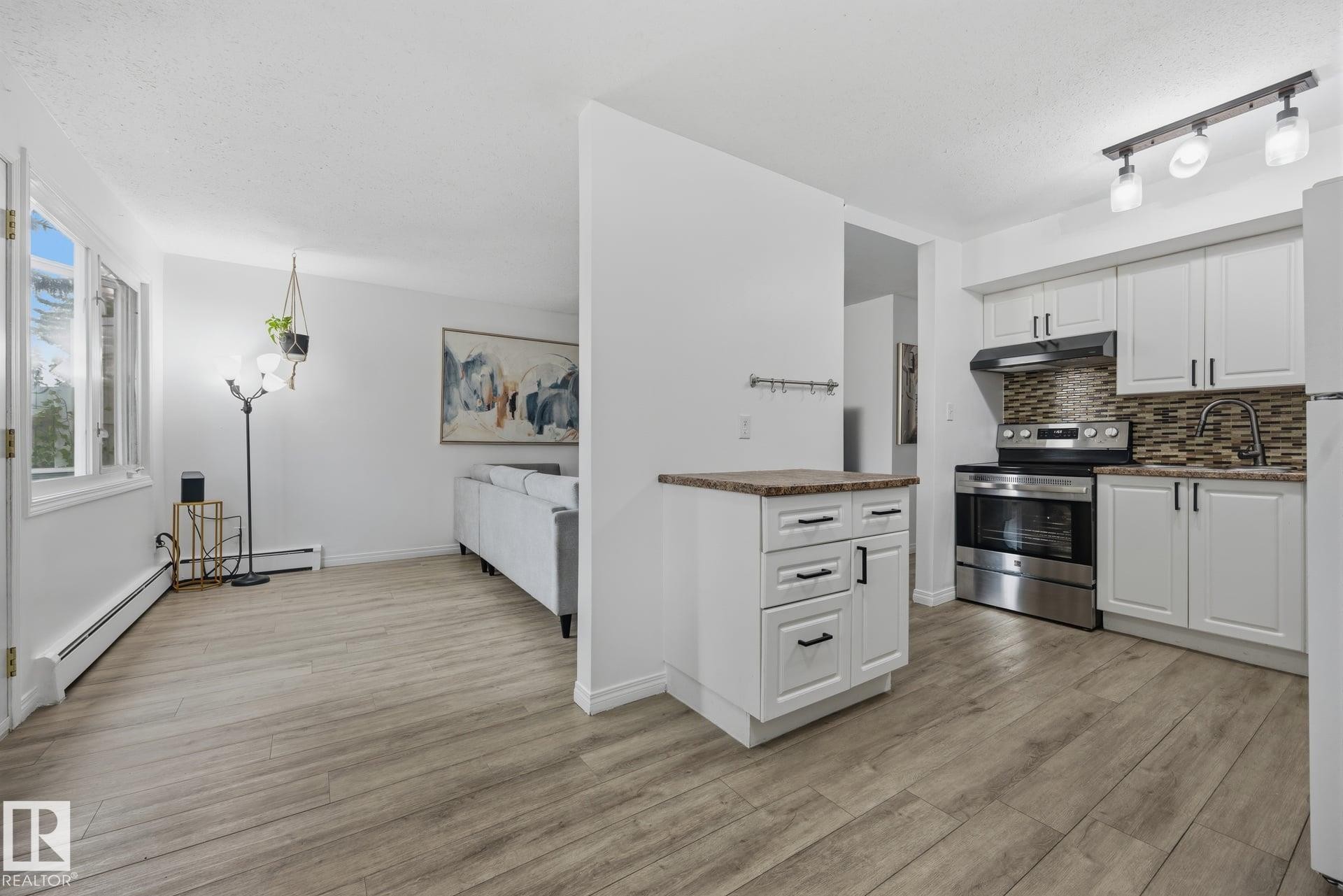 Open-concept living area featuring wood-finish flooring, white cabinetry, stainless steel appliances, a mosaic tile backsplash, and a functional half-wall island - 105 16428 109 Street, Edmonton, AB