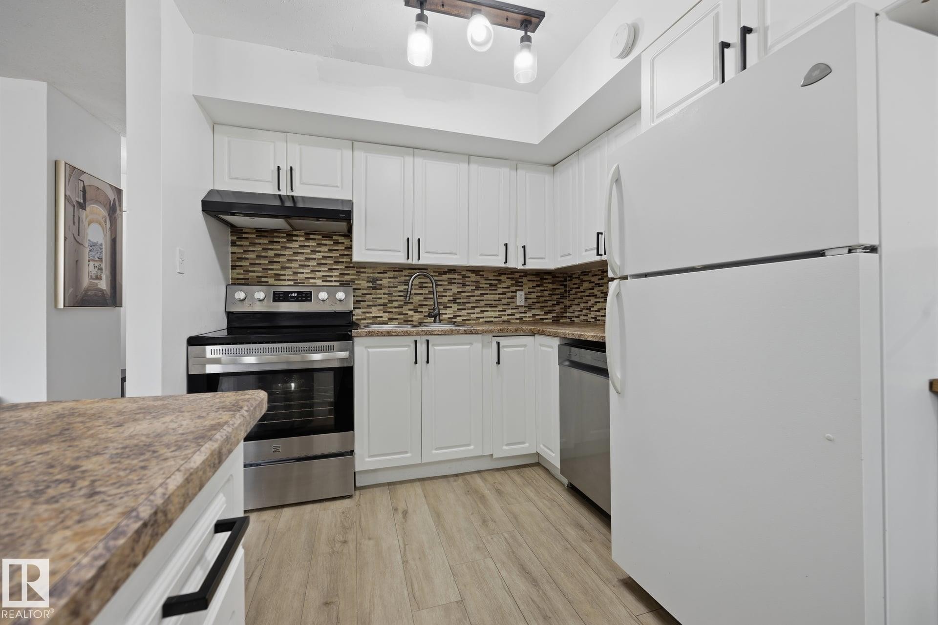 Galley kitchen featuring white cabinetry with matte black hardware, stainless steel appliances, mosaic tile backsplash, wood-finish flooring, and a peninsula with a stone-look countertop - 105 16428 109 Street, Edmonton, AB