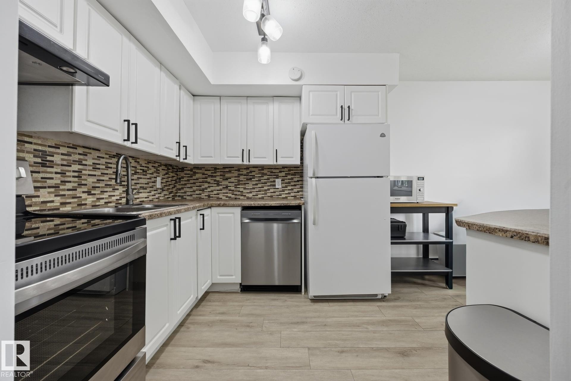 Modern kitchen featuring white cabinetry, a mosaic tile backsplash, wood-finish flooring, and stainless steel appliances including an oven range and dishwasher - 105 16428 109 Street, Edmonton, AB