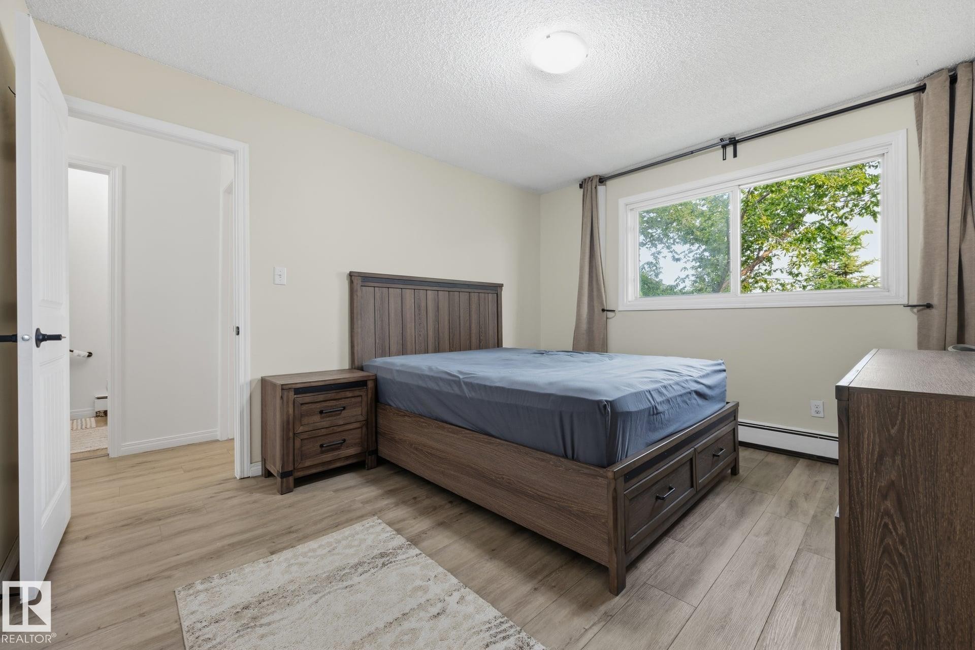 Bedroom featuring light wood-finish flooring, a bright window with neutral drapes, and a white ceiling with a flush-mount light fixture - 105 16428 109 Street, Edmonton, AB