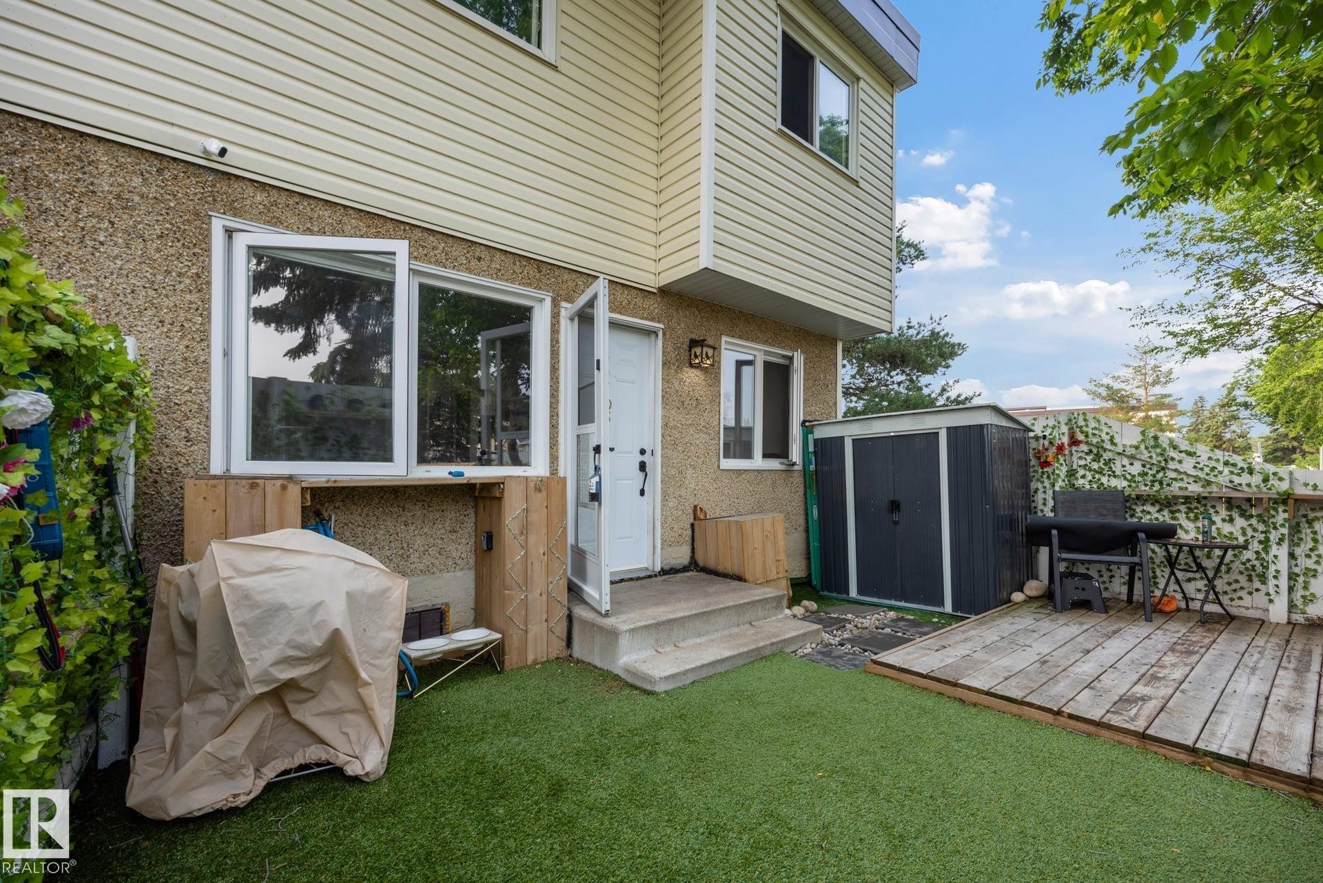 Rear exterior featuring a ground-level patio door and windows, artificial turf, wooden decking, a detached storage shed, and a privacy fence - 105 16428 109 Street, Edmonton, AB
