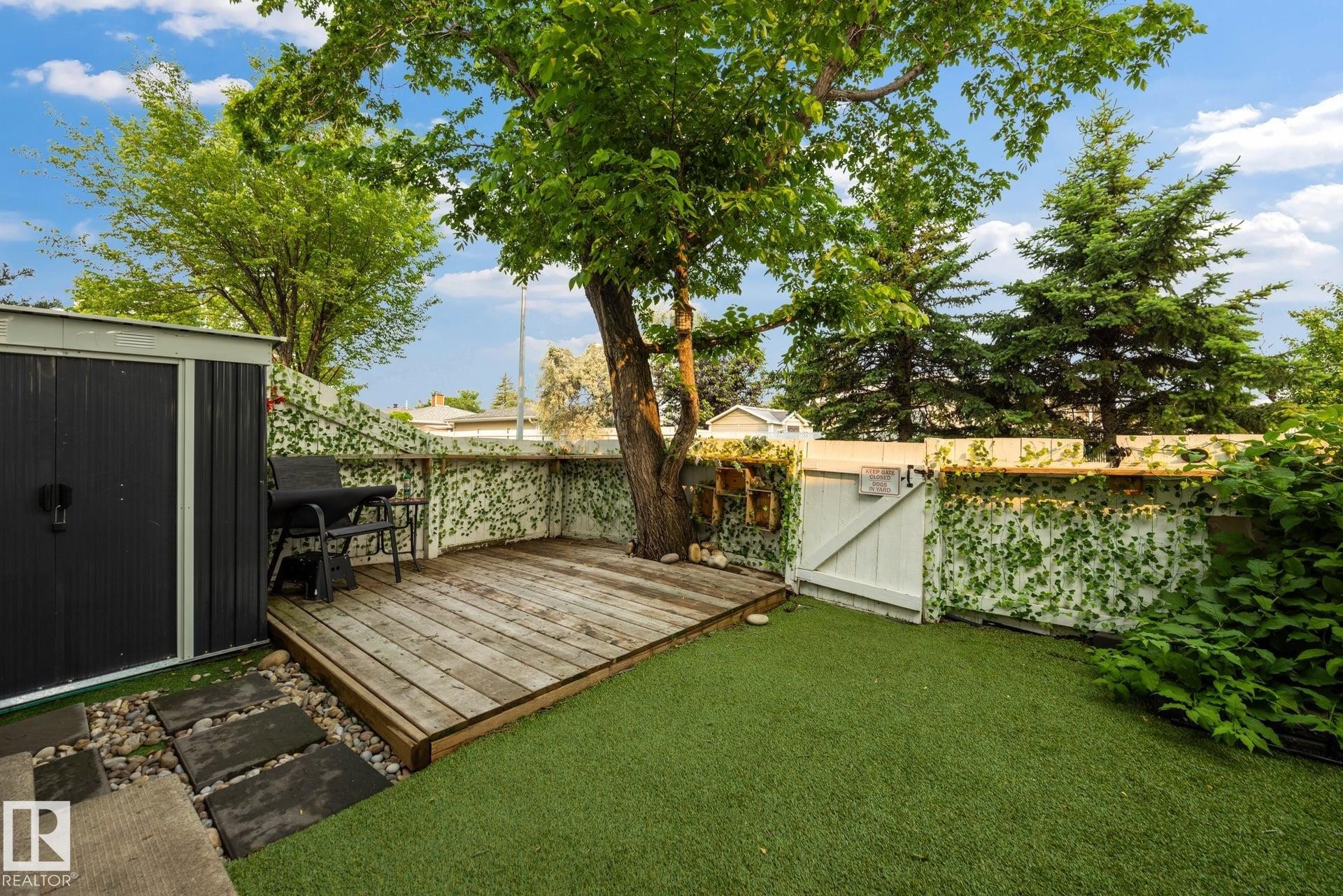 Backyard featuring a wood-finish deck, artificial turf, stepping stones, and a storage shed - 105 16428 109 Street, Edmonton, AB