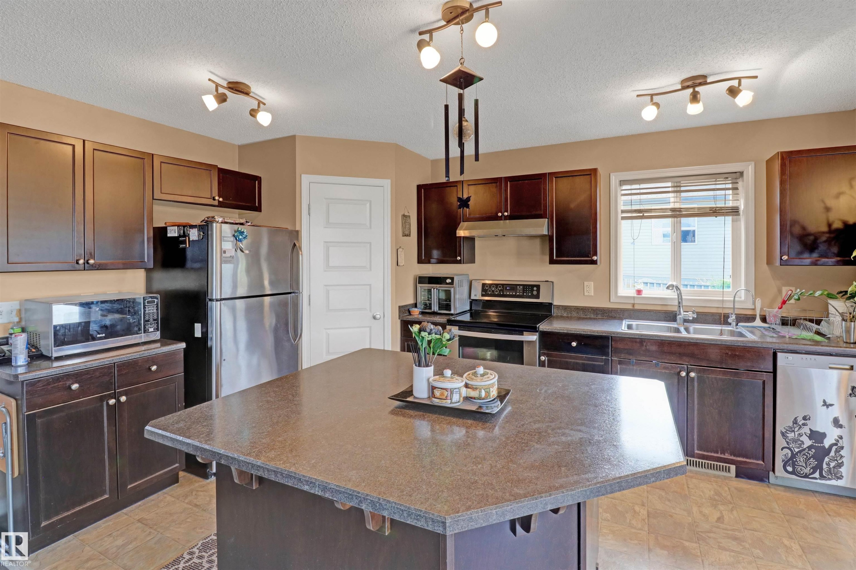 Kitchen featuring a central island with a speckled countertop, dark wood-finish cabinetry, stainless steel appliances, and track lighting fixtures - 1216 37C Avenue Nw, Edmonton, AB - Indoor Photo Showing Kitchen With Double Sink