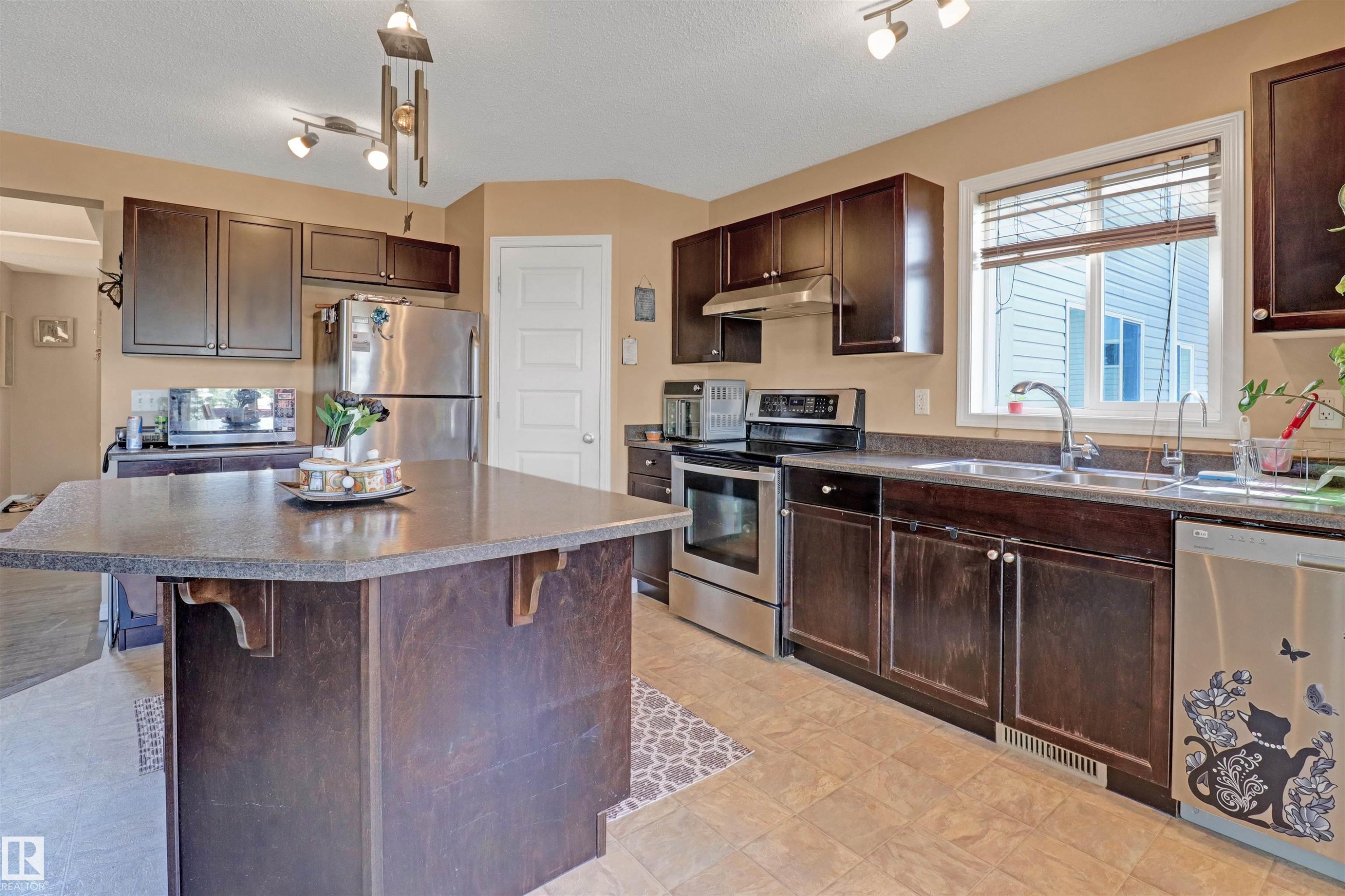 Kitchen featuring dark wood-finish cabinetry, stainless steel appliances, a large island with an extended countertop, and light-toned flooring - 1216 37C Avenue Nw, Edmonton, AB - Indoor Photo Showing Kitchen With Double Sink