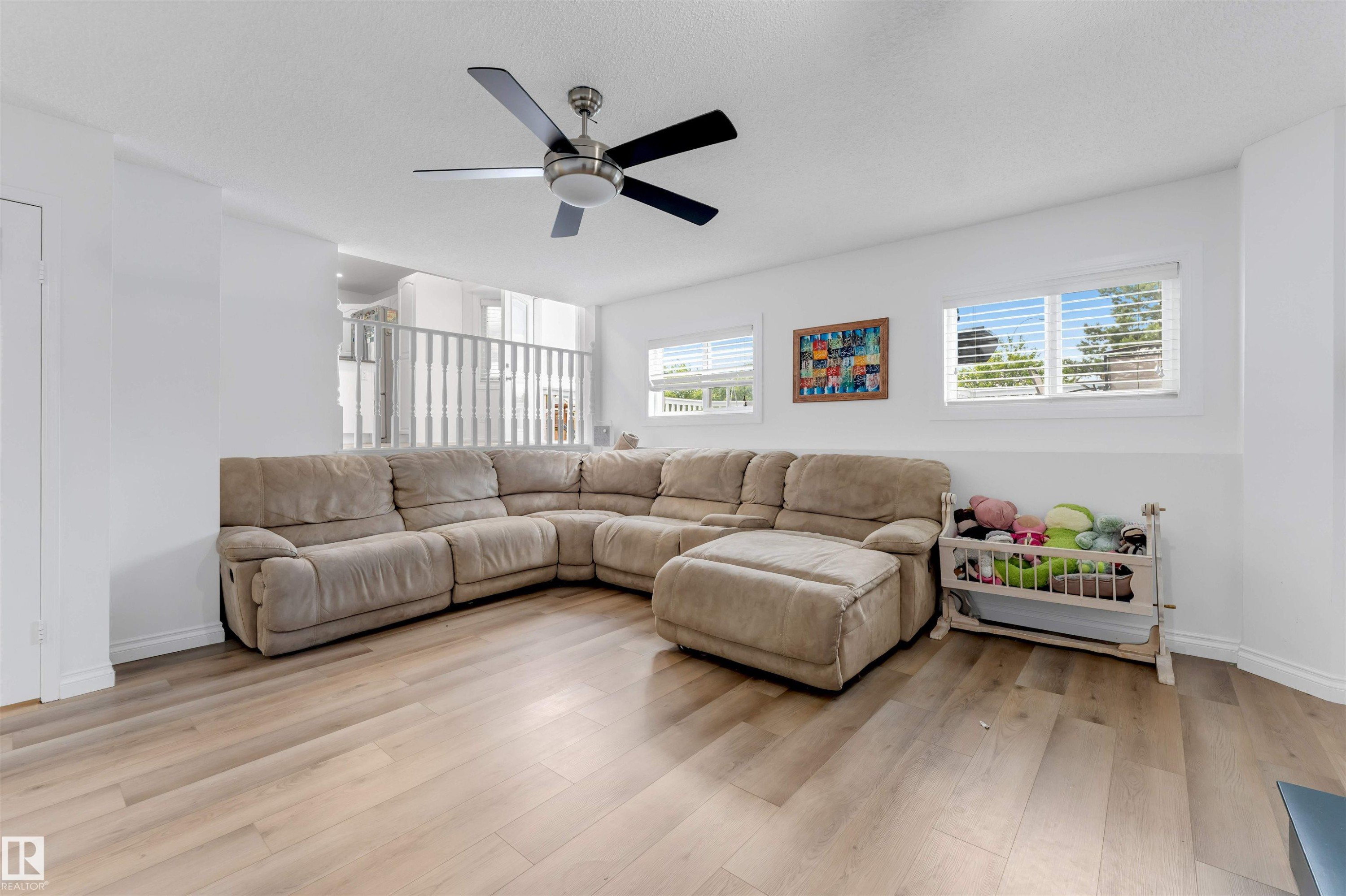 Spacious room featuring light wood-finish flooring, multiple windows with blinds, a ceiling fan with integrated lighting, and an open-concept railing overlooking another space - 3332 43 Street Nw, Edmonton, AB