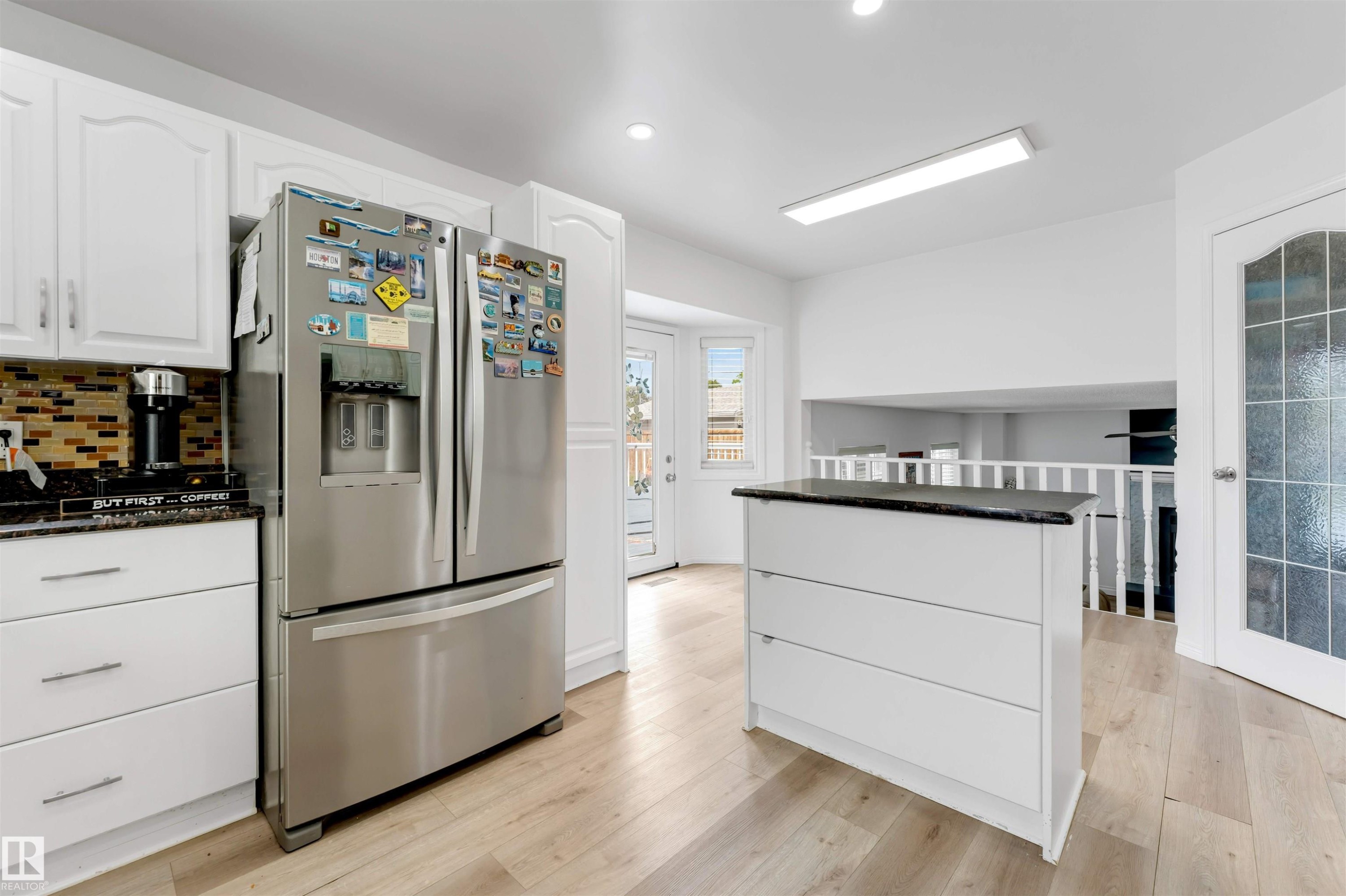 Bright kitchen featuring white cabinetry, light wood-finish flooring, a mosaic tile backsplash, a central island with a dark countertop, and a glass-paneled door - 3332 43 Street Nw, Edmonton, AB