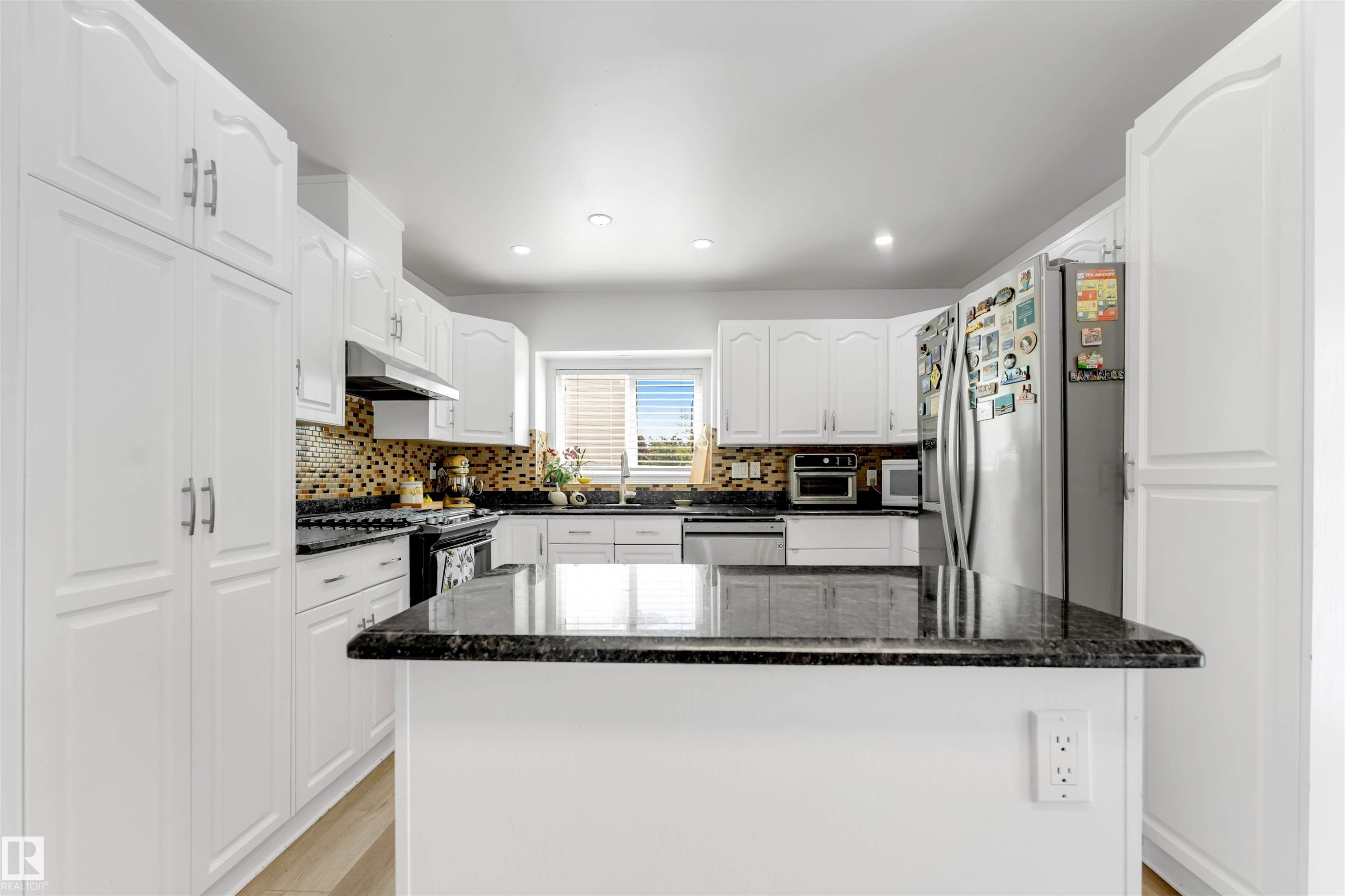 Kitchen featuring white cabinetry, dark stone countertops, stainless steel appliances, a mosaic tile backsplash, and a central island - 3332 43 Street Nw, Edmonton, AB