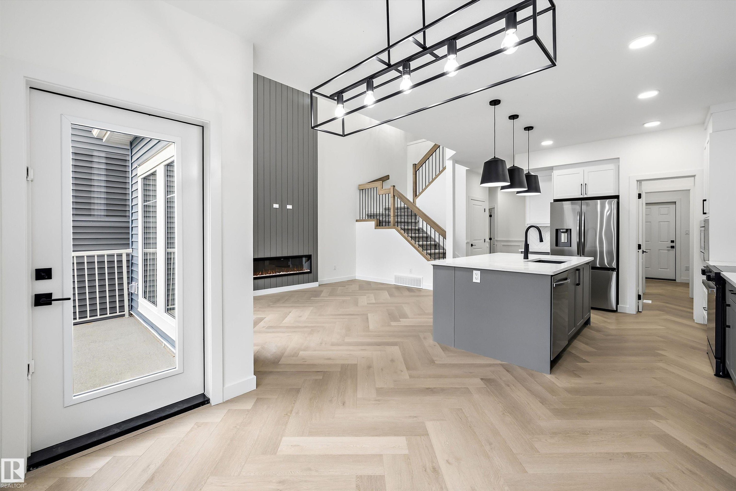 Open-concept living space featuring wood-finish herringbone flooring, a contemporary kitchen island with a white countertop and dark pendant lighting, and a modern linear fireplace - 917 Elderberry Landing, Edmonton, AB