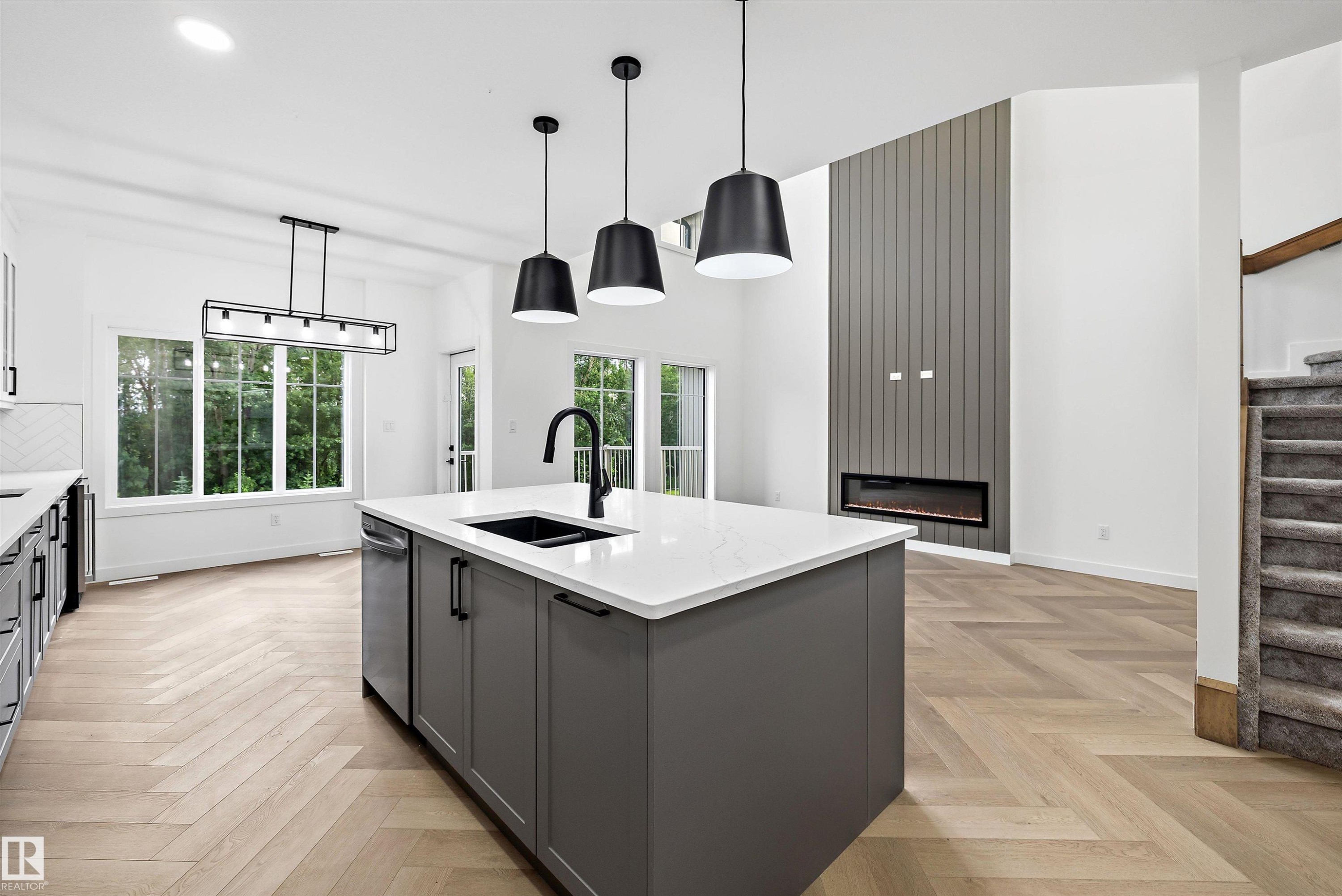 Spacious open-concept living area featuring a kitchen island with stone countertop and integrated sink, chevron wood-finish flooring, a contemporary linear fireplace with a vertical panel accent wall, and large windows providing abundant natural light - 917 Elderberry Landing, Edmonton, AB