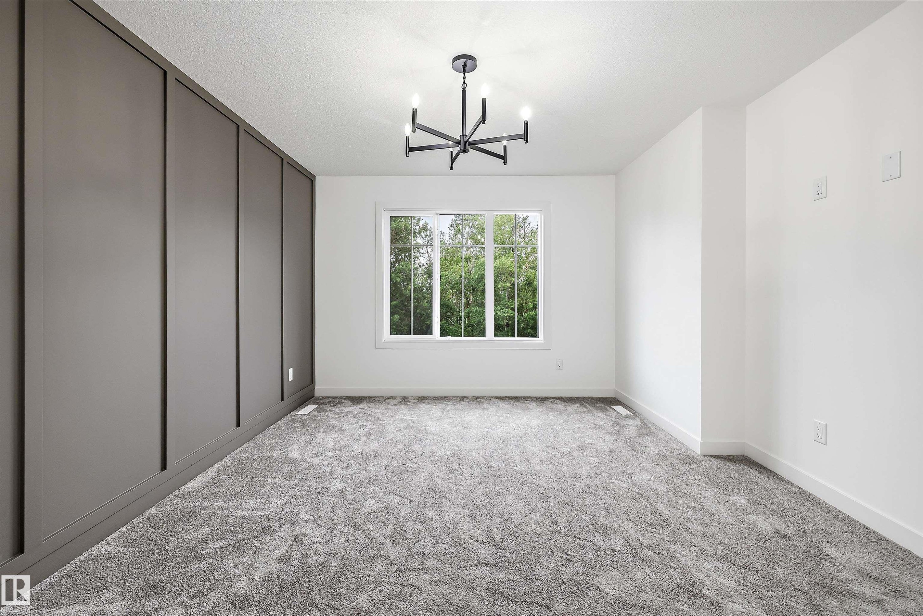 Room featuring a modern black chandelier, a large white-framed window, textured gray carpeting, a paneled accent wall, and clean white walls - 917 Elderberry Landing, Edmonton, AB