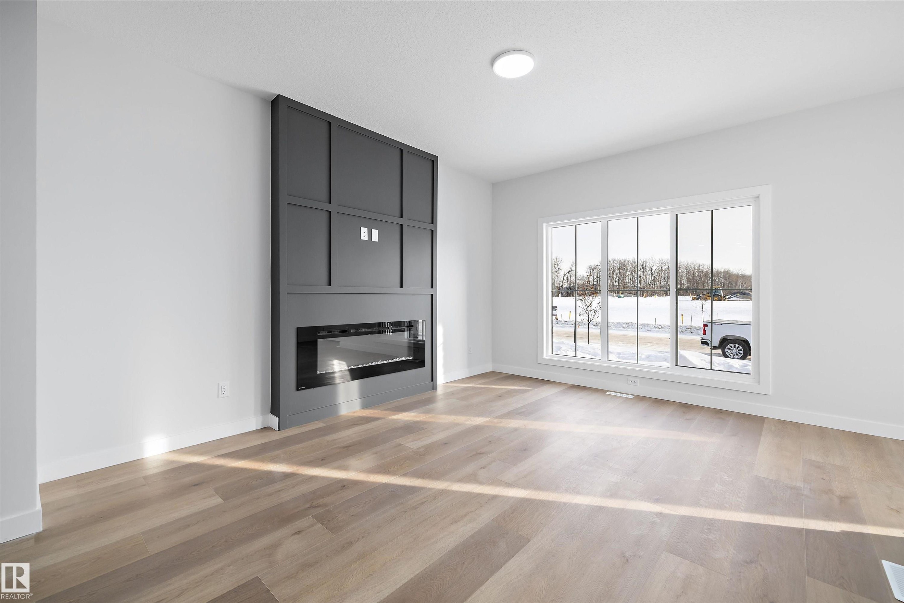 Spacious living area featuring a dark gray accent wall with an integrated linear fireplace, large picture window, light wood-finish flooring, and recessed ceiling lighting - 954 Elderberry Landing, Edmonton, AB