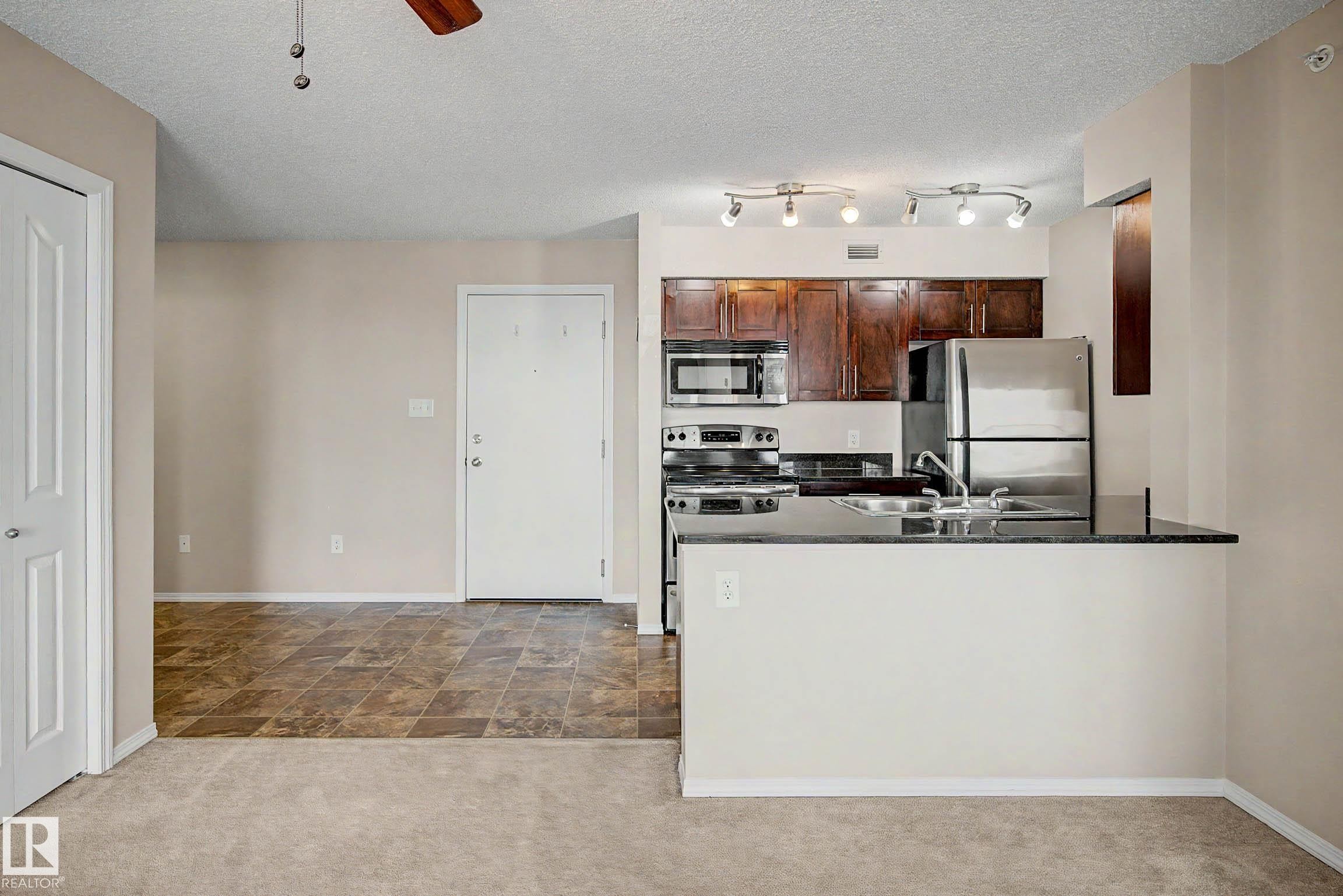 Open-concept kitchen featuring dark wood cabinetry, stainless steel appliances, a black countertop, and a breakfast bar - 404 1080 Mcconachie Boulevard, Edmonton, AB