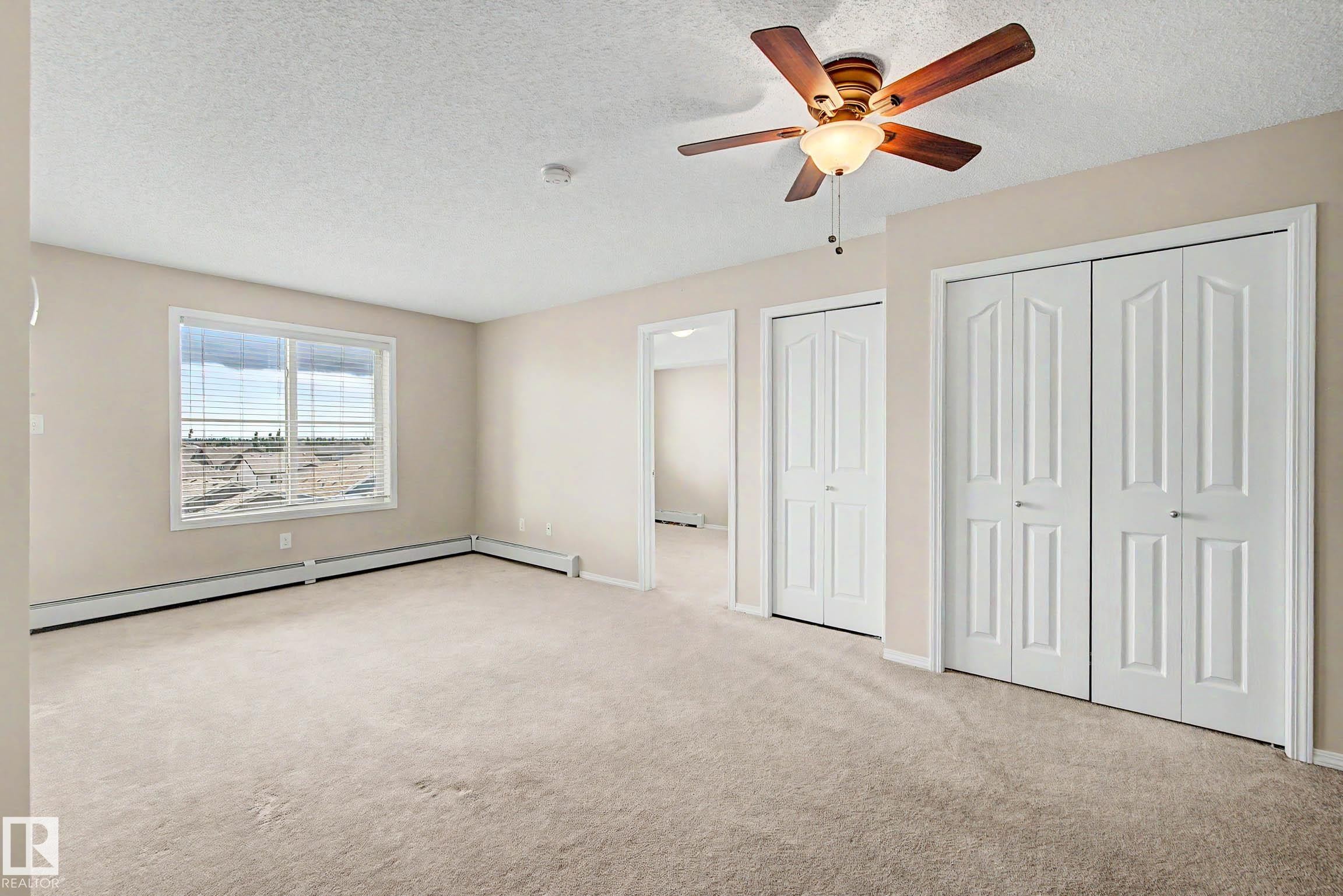 Spacious room featuring neutral carpet flooring, a window offering exterior views, two bi-fold panel doors, and a ceiling fan with integrated lighting - 404 1080 Mcconachie Boulevard, Edmonton, AB