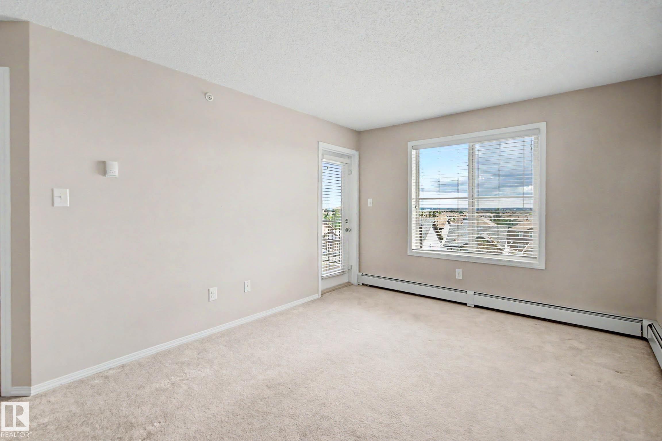 Spacious interior featuring neutral wall tones, textured carpet flooring, and a large window with horizontal blinds - 404 1080 Mcconachie Boulevard, Edmonton, AB