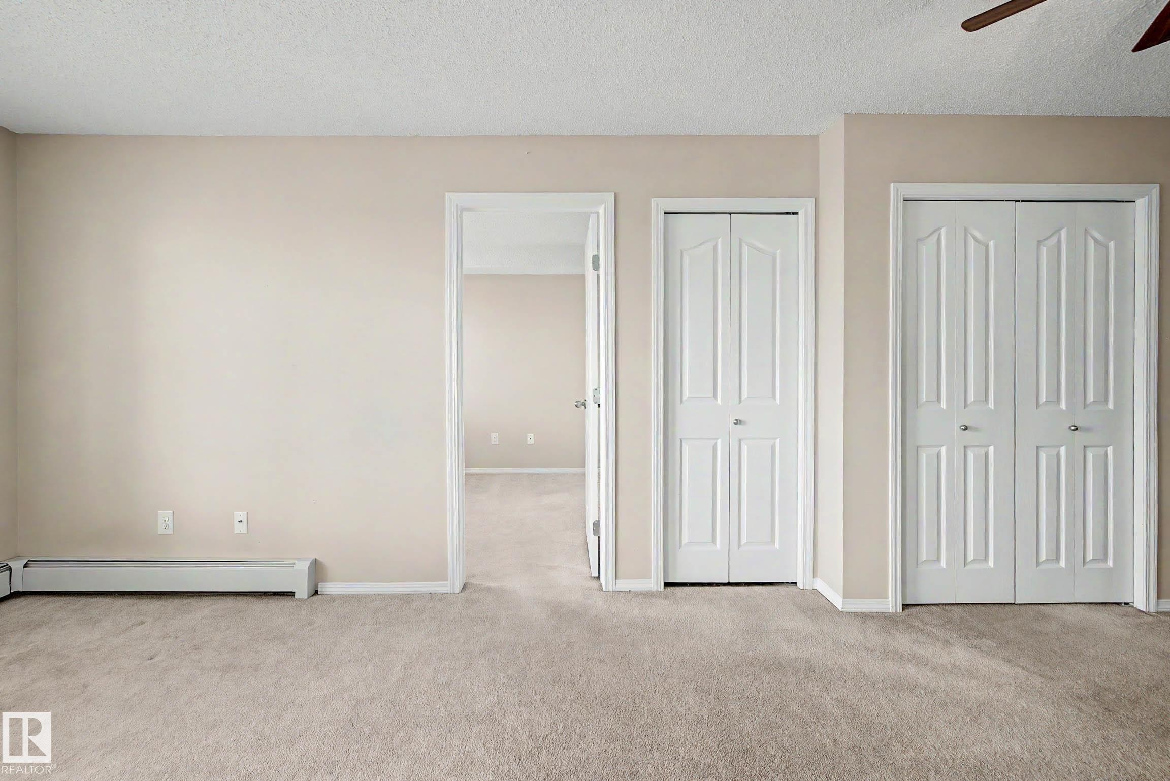 Carpeted interior space featuring neutral wall paint, a white baseboard heater, and multiple white bi-fold doors - 404 1080 Mcconachie Boulevard, Edmonton, AB