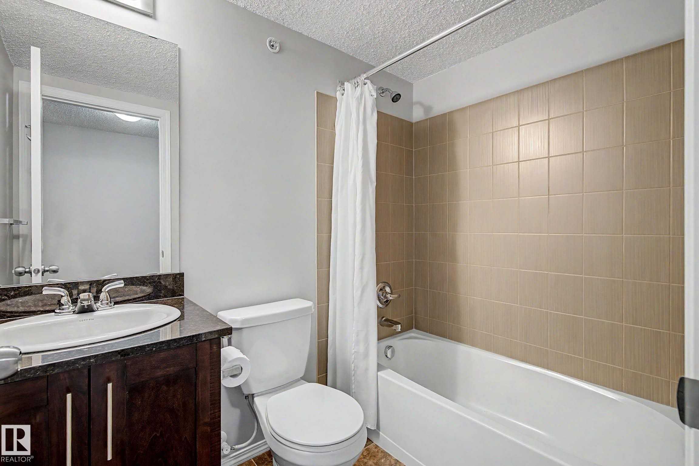 Bathroom featuring a dark wood-finish vanity with a stone countertop and undermount sink, a toilet, a bathtub with a shower, and beige tile surround - 404 1080 Mcconachie Boulevard, Edmonton, AB