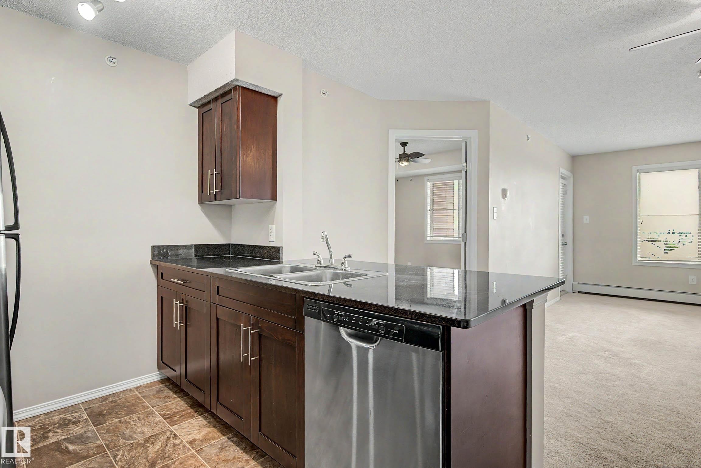 Kitchen featuring dark wood-finish cabinetry, a stainless steel double sink, and a stainless steel dishwasher - 404 1080 Mcconachie Boulevard, Edmonton, AB