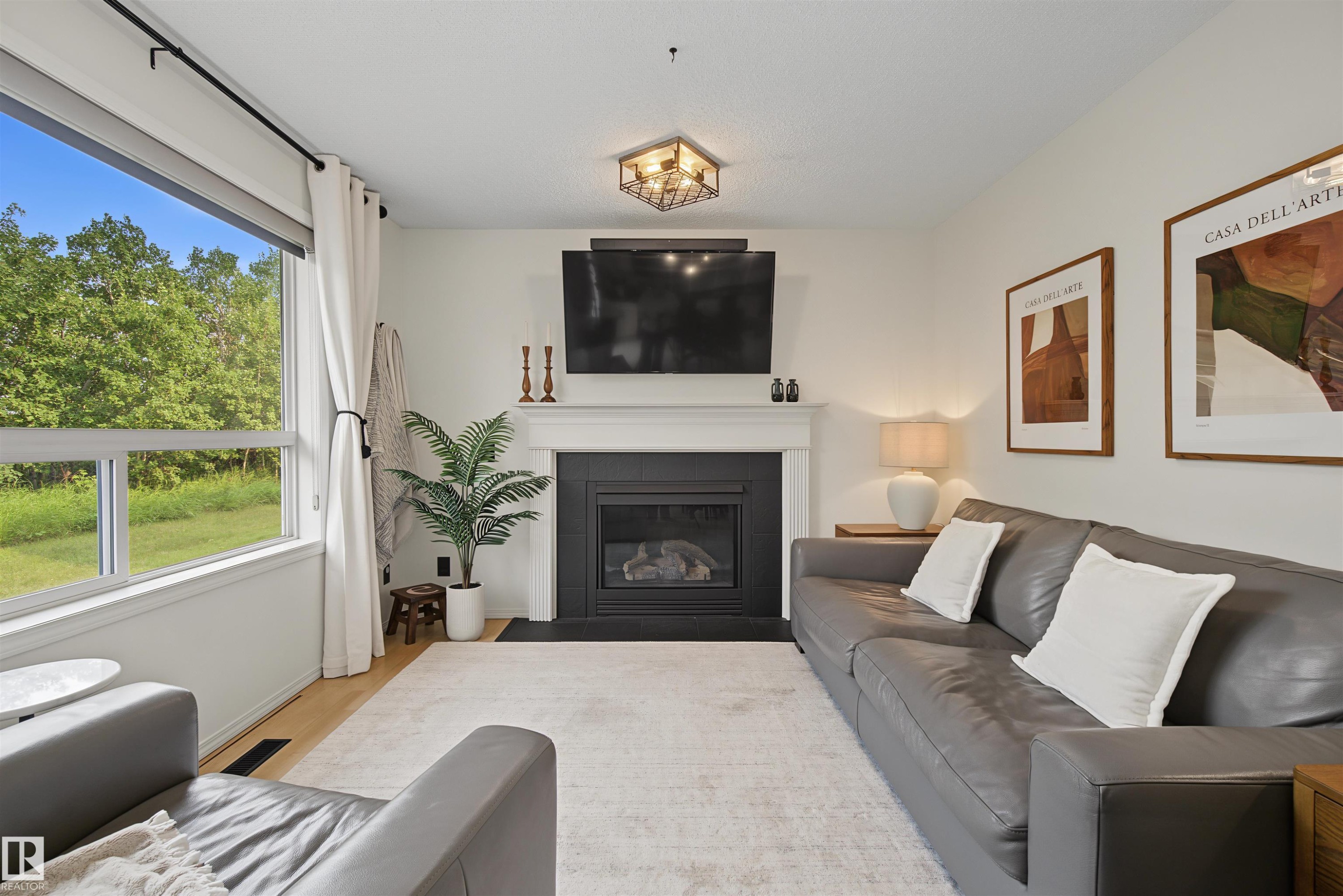 Living room featuring a large window, light wood-finish flooring, a fireplace with white mantel, and a ceiling-mounted light fixture - 15 450 Hyndman Crescent, Edmonton, AB