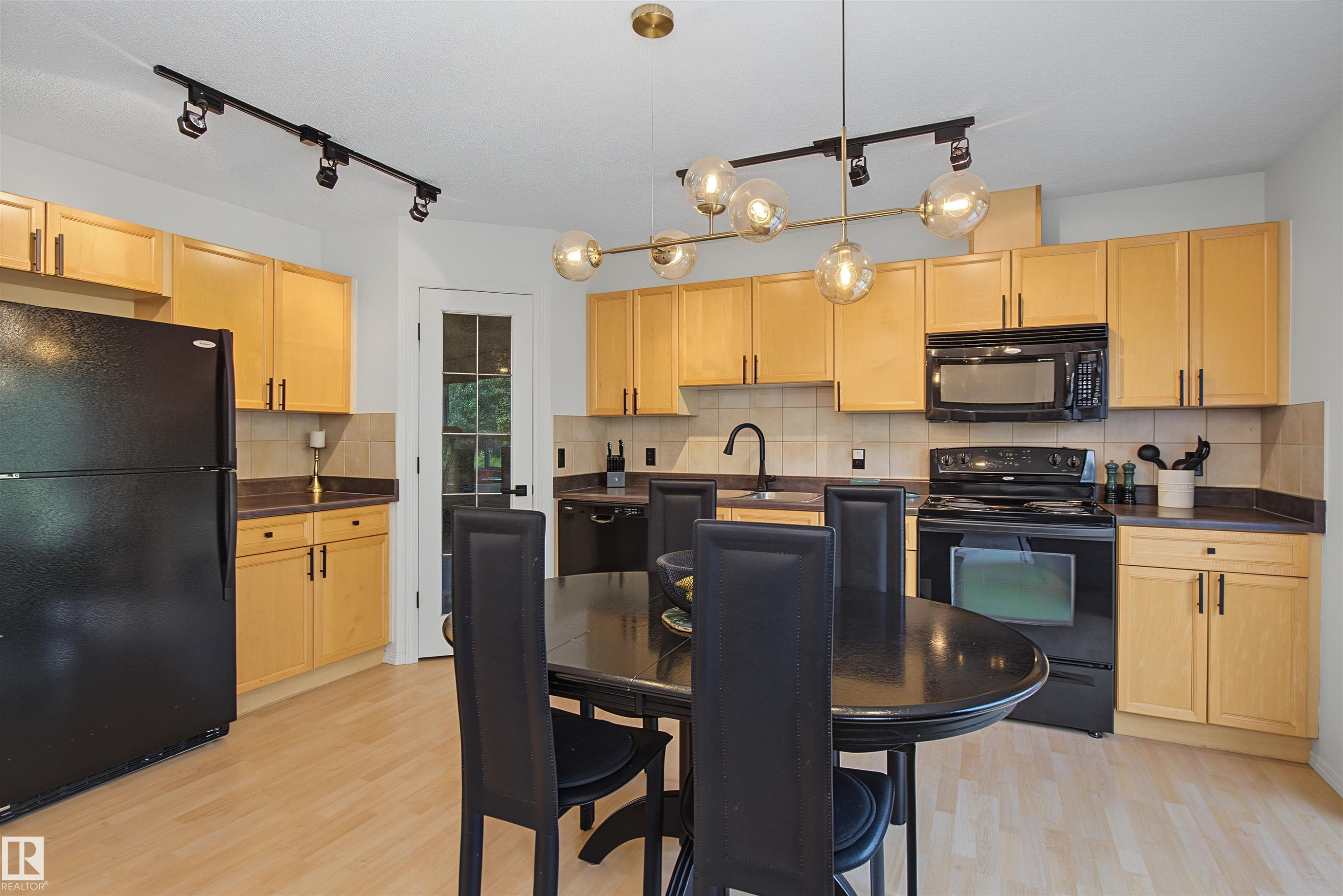 Kitchen featuring light wood-finish cabinetry, black appliances, a tile backsplash, and wood-finish flooring - 15 450 Hyndman Crescent, Edmonton, AB