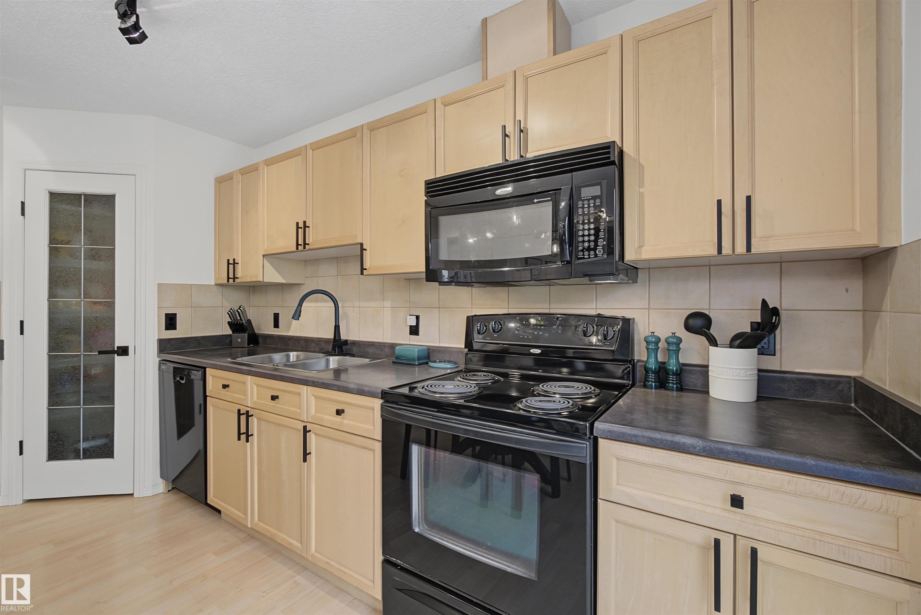 Kitchen featuring light wood-finish cabinetry with black hardware, black appliances, solid surface countertops, and a tile backsplash - 15 450 Hyndman Crescent, Edmonton, AB