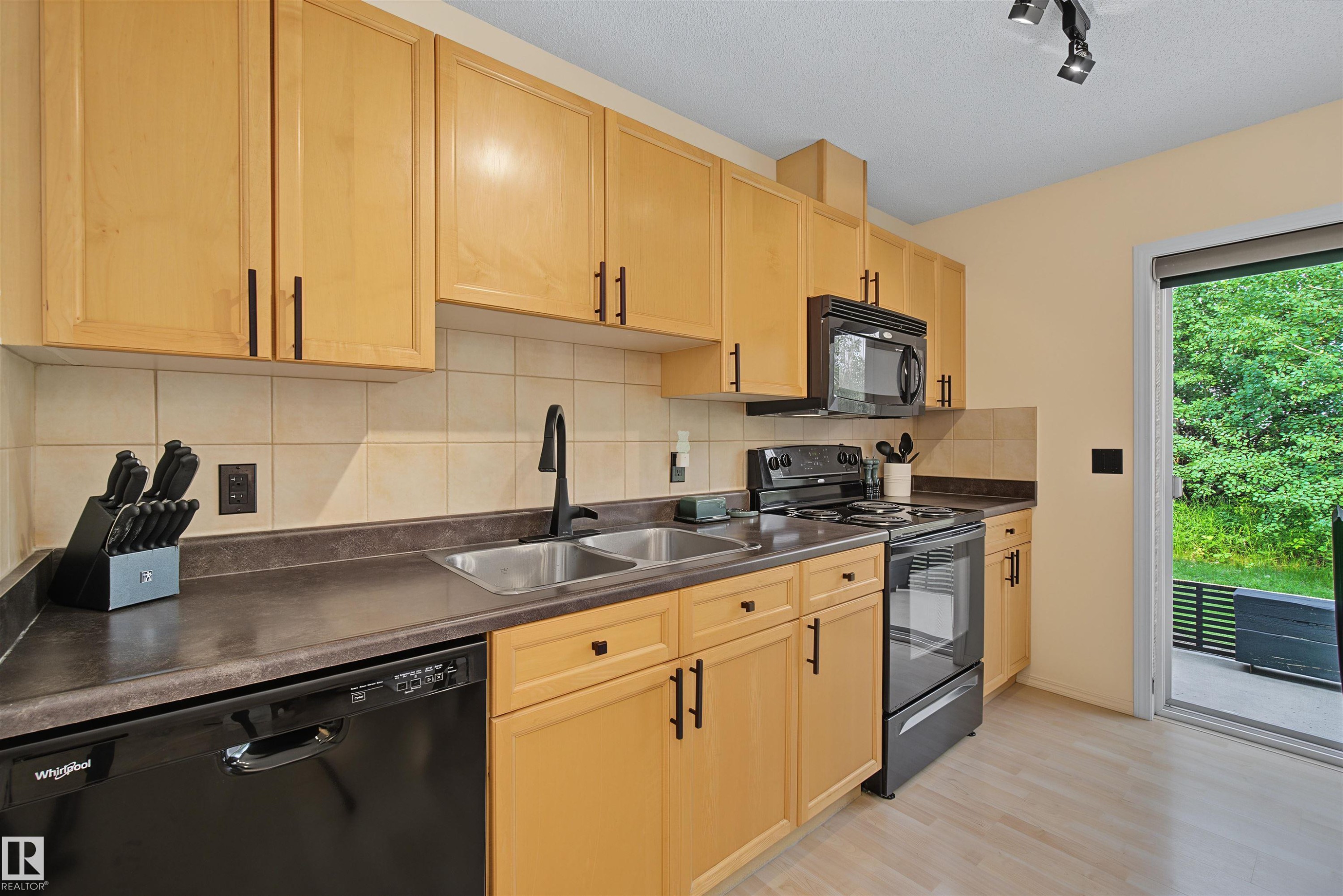 Kitchen featuring light wood cabinetry with dark hardware, black appliances, a tile backsplash, and dark countertops - 15 450 Hyndman Crescent, Edmonton, AB