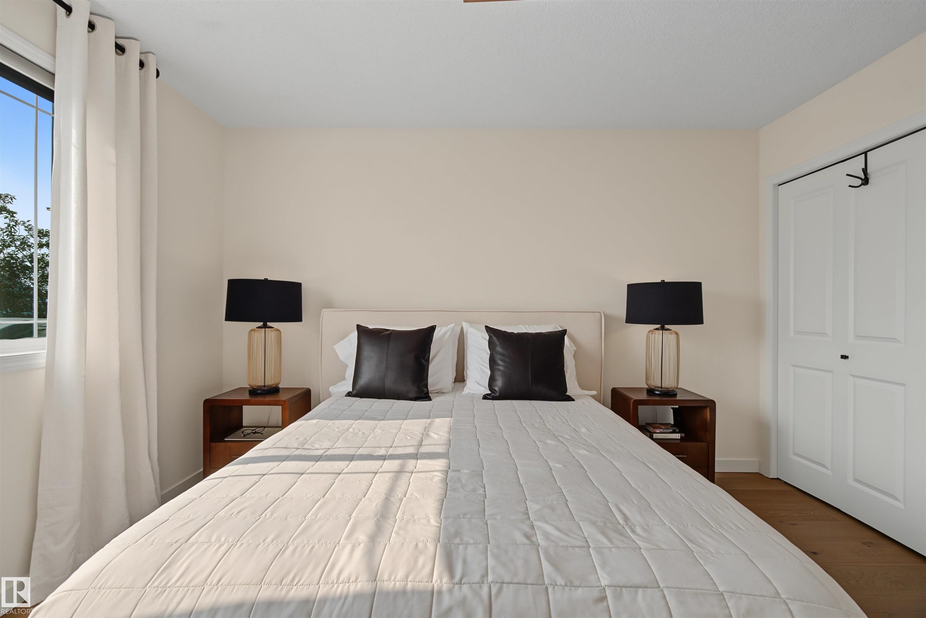 Bedroom featuring a large window with white curtains, light-toned walls, and wood-finish flooring - 15 450 Hyndman Crescent, Edmonton, AB