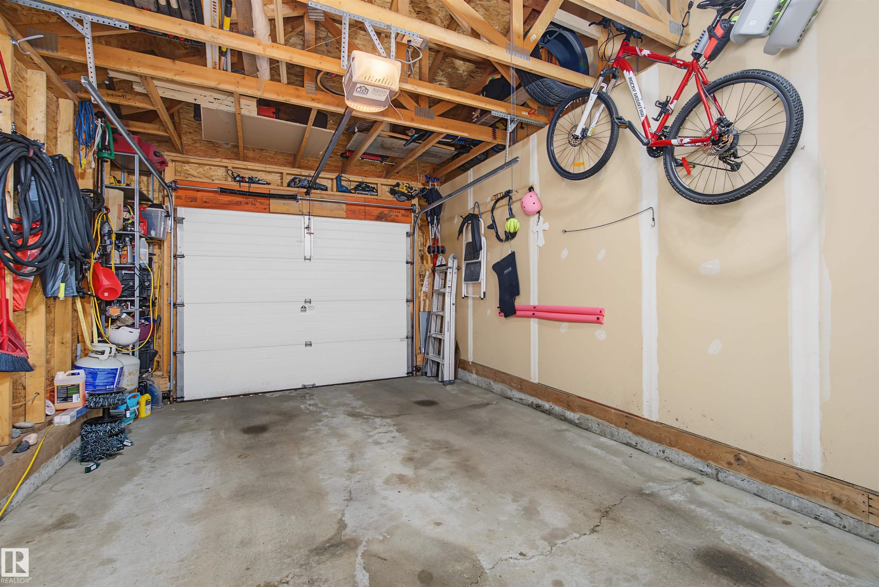 Single-bay garage featuring a concrete floor, exposed wood ceiling joists, and a white panel garage door with an automatic opener - 15 450 Hyndman Crescent, Edmonton, AB
