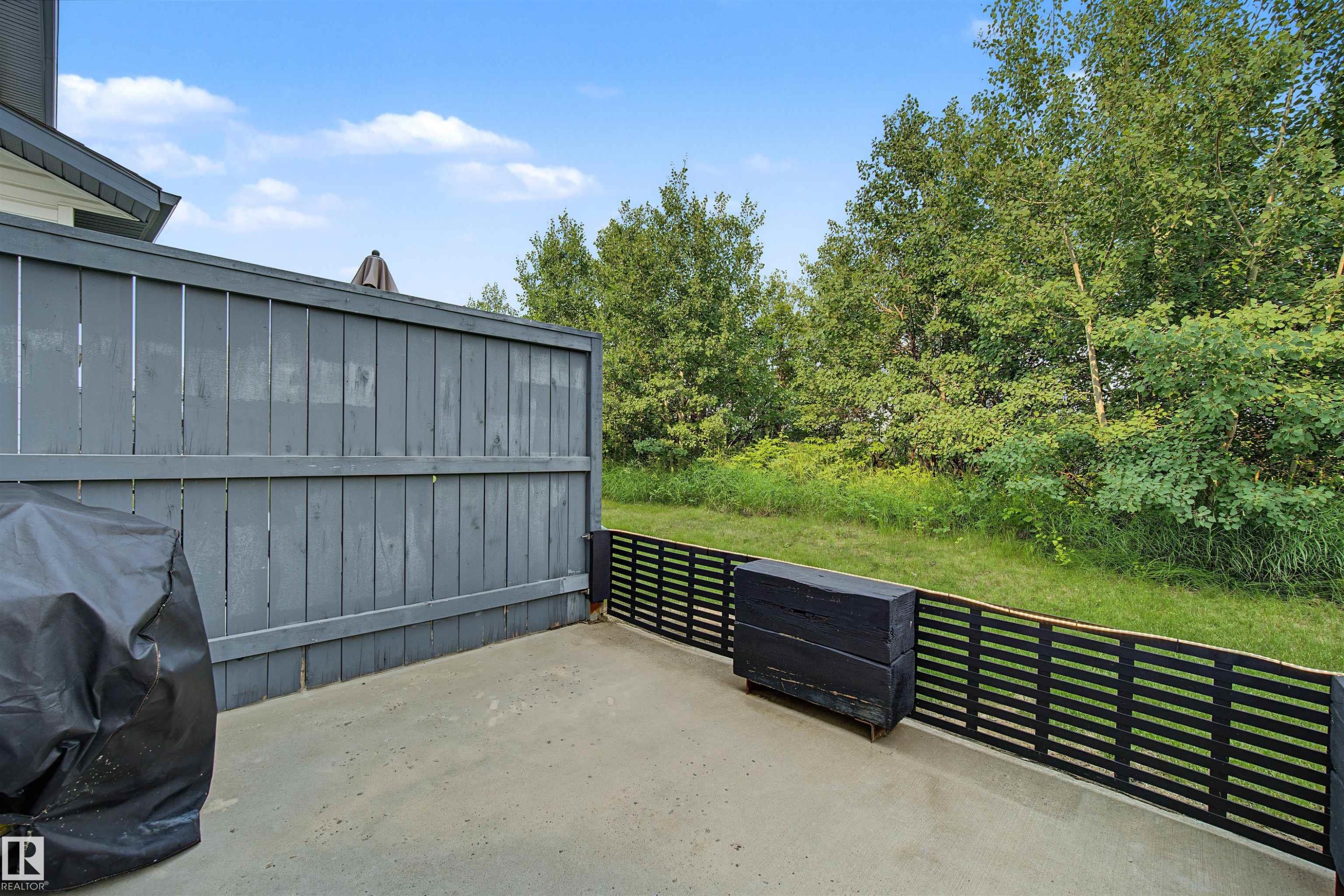 Outdoor patio with concrete flooring, a partial privacy fence, and a horizontal slat perimeter fence, bordered by mature trees - 15 450 Hyndman Crescent, Edmonton, AB