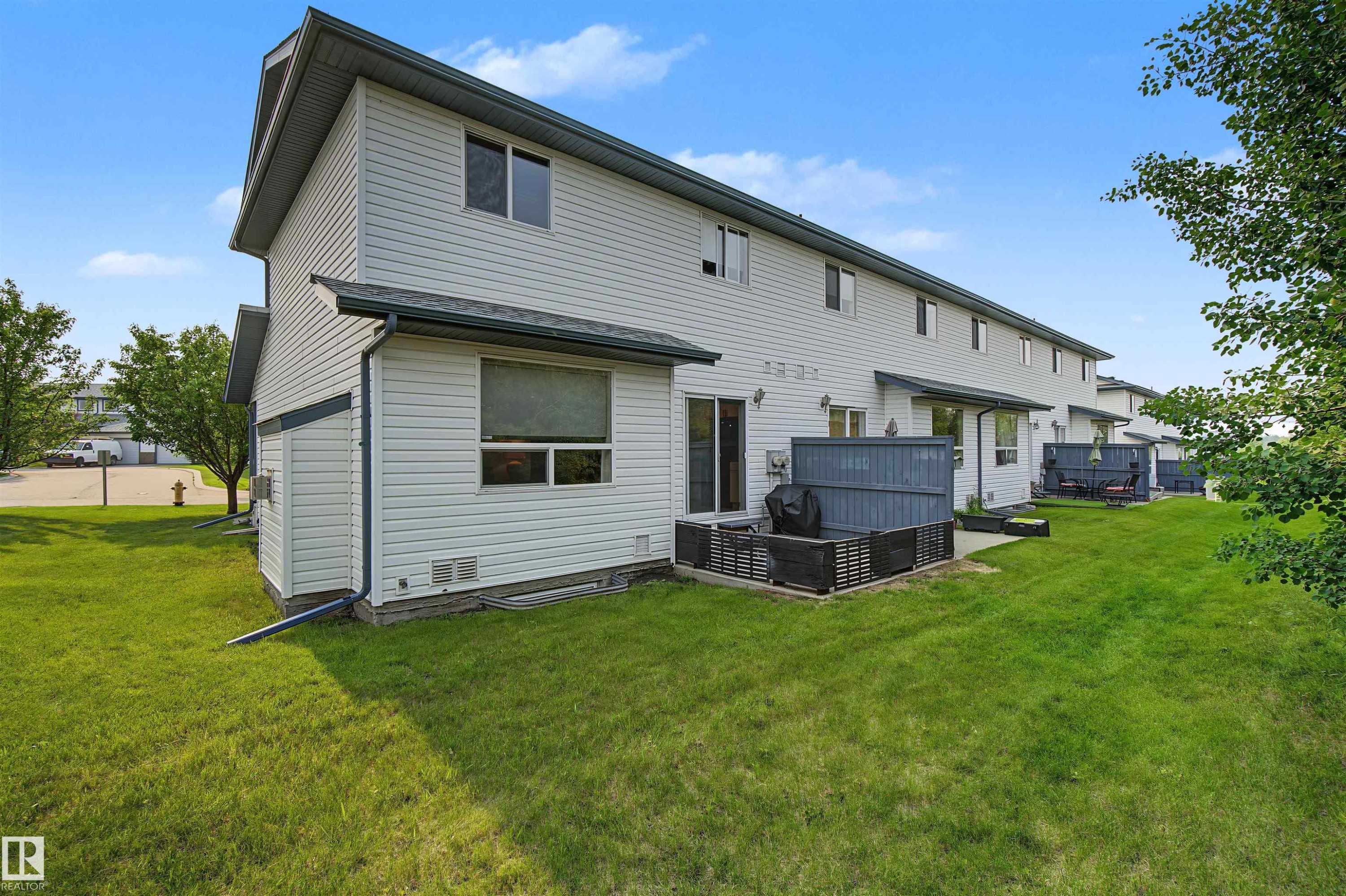 Rear exterior showcasing white siding, multiple windows, and a sliding glass door leading to a private patio area - 15 450 Hyndman Crescent, Edmonton, AB