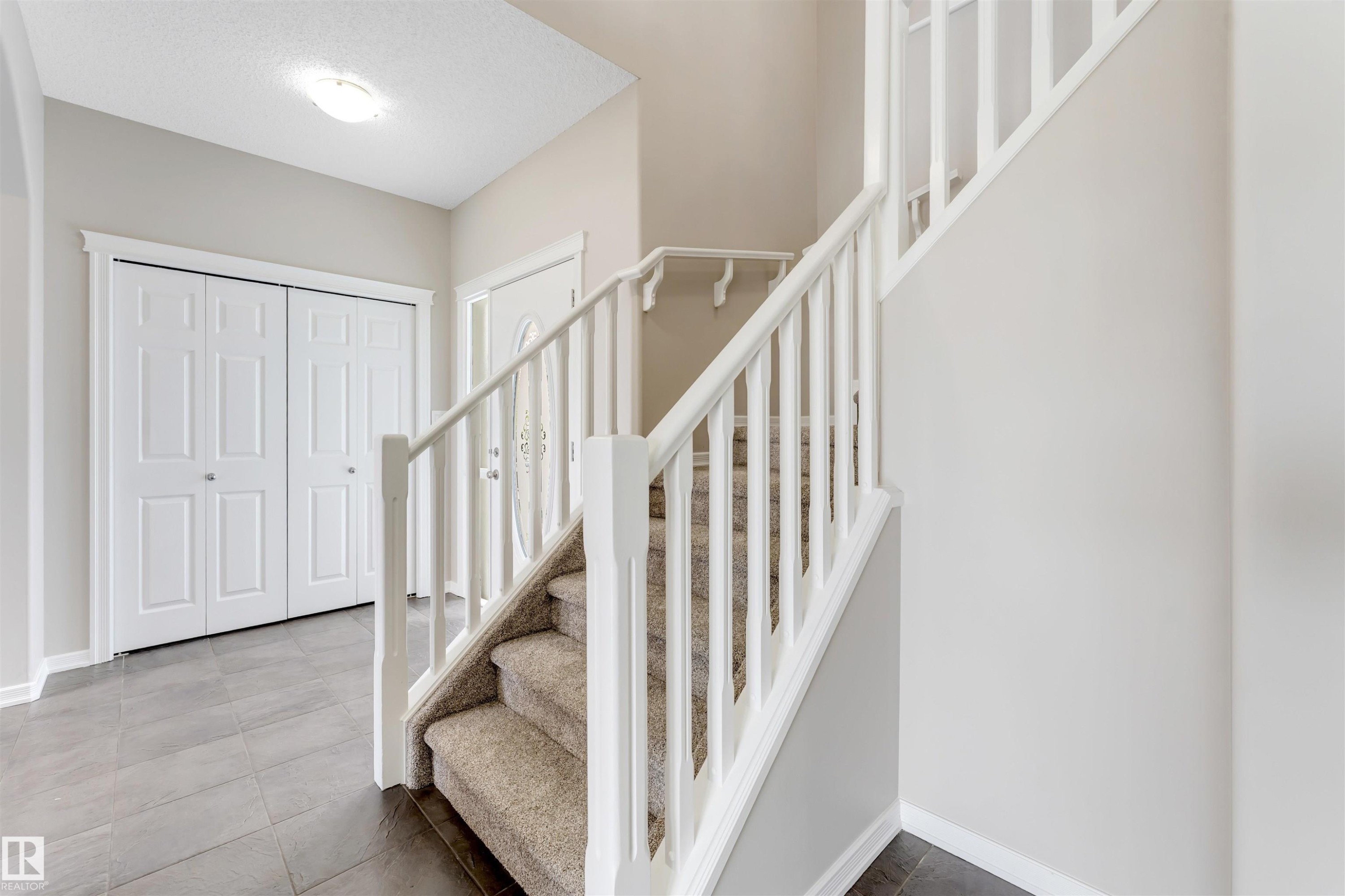 Entryway featuring a carpeted staircase with white balusters, gray floor tiles, bi-fold closet doors, and a front door with decorative glass - 3449 28 Street, Edmonton, AB