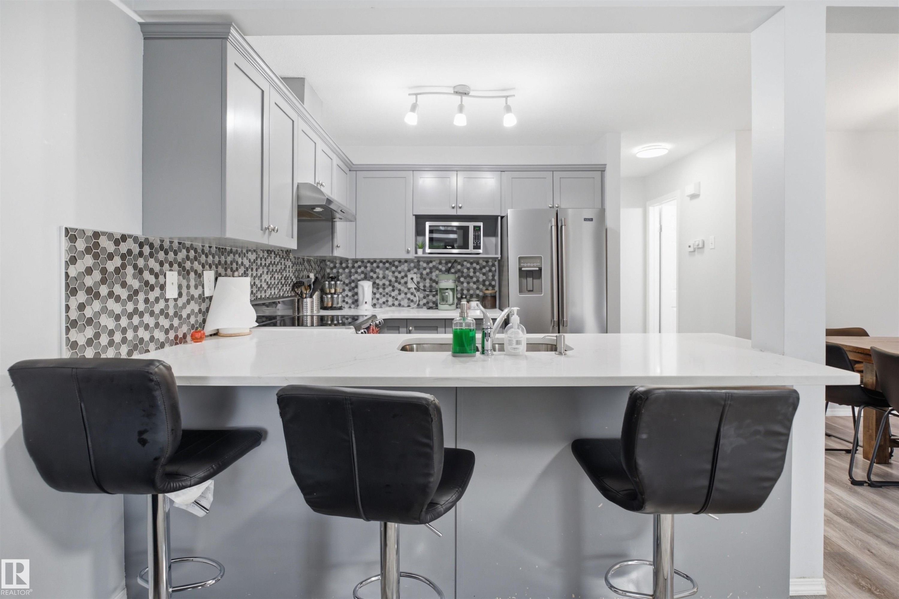 Modern kitchen featuring a breakfast bar with a white countertop, gray flat-panel cabinetry, a mosaic tile backsplash, and stainless steel appliances - 92 230 Edwards Drive, Edmonton, AB
