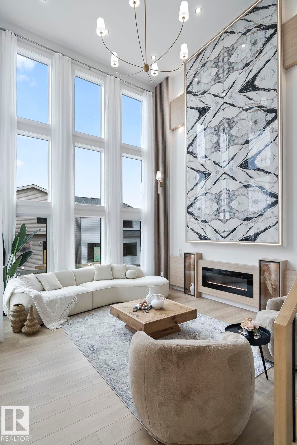 Double-height living space featuring expansive windows with white drapery, wood-finish flooring, a contemporary chandelier, a built-in electric fireplace, and a large statement art piece - 13152 187 Avenue, Edmonton, AB