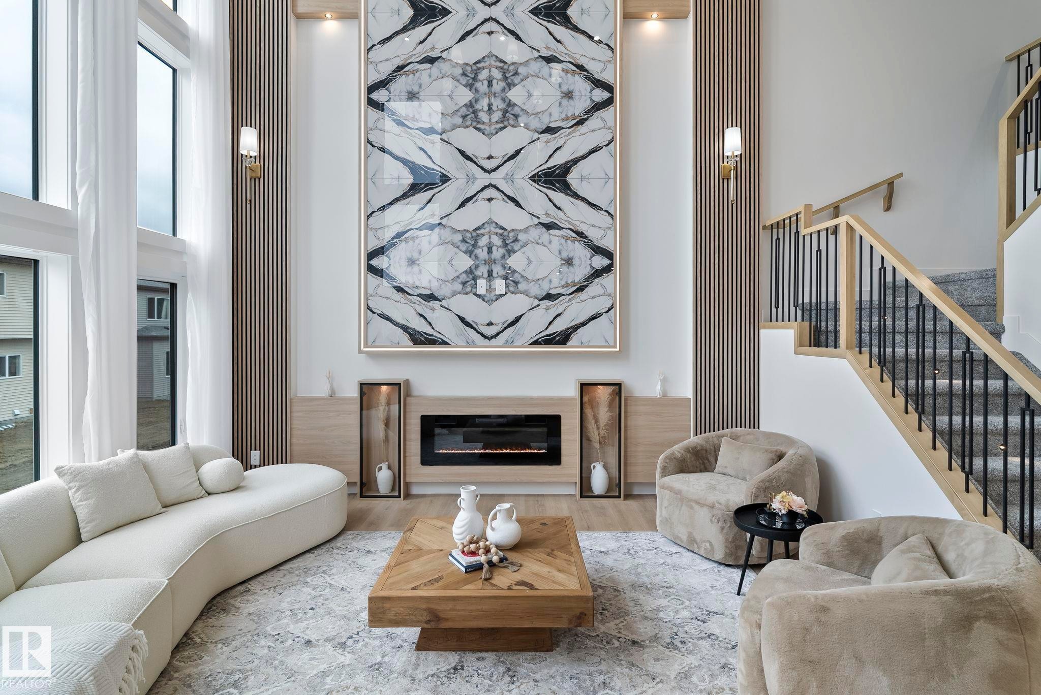 Double-height living space featuring expansive windows, a built-in electric fireplace, wood-finish wall paneling, and an open staircase with metal balusters - 13152 187 Avenue, Edmonton, AB