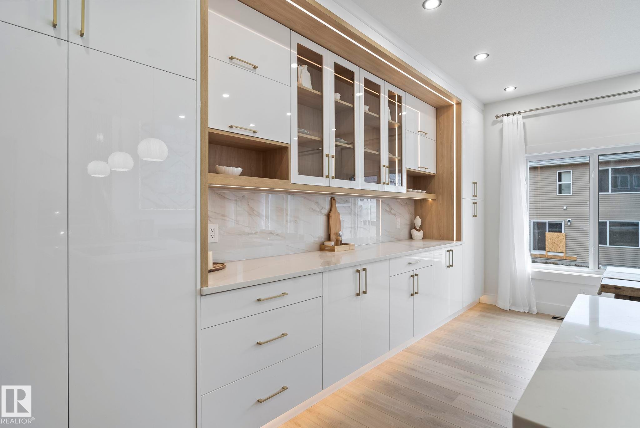 Integrated white cabinetry with gold-tone hardware, wood-finish shelving, glass-front upper cabinets, and a white countertop with a complementing backsplash - 13152 187 Avenue, Edmonton, AB