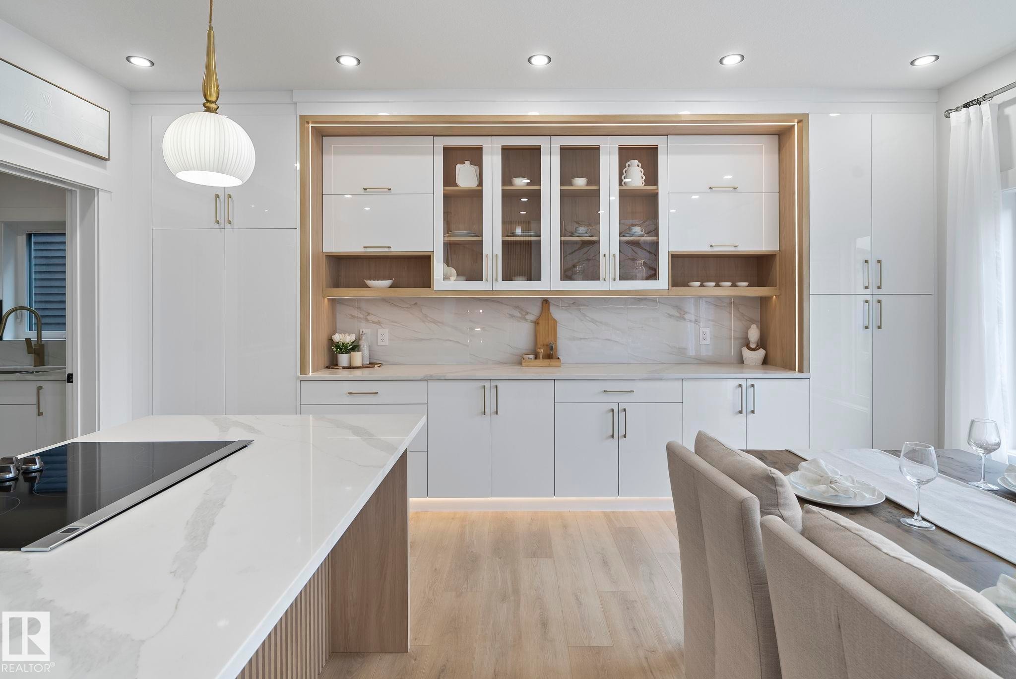 Modern kitchen featuring an island with a built-in cooktop, white shaker-style cabinetry with glass-front uppers, light wood-finish flooring, recessed lighting, and a white pendant light fixture - 13152 187 Avenue, Edmonton, AB