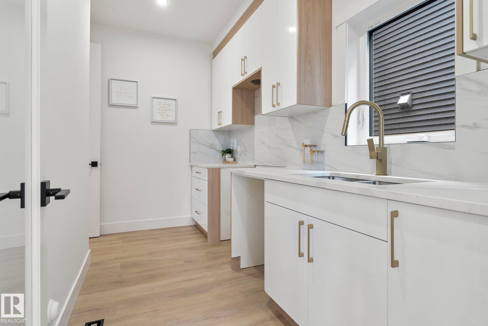 Contemporary laundry area featuring a stainless steel sink with a brass-finish faucet, quartz-finish countertops, full-height tile backsplash, white and wood-finish cabinetry, and wood-finish flooring - 13152 187 Avenue, Edmonton, AB