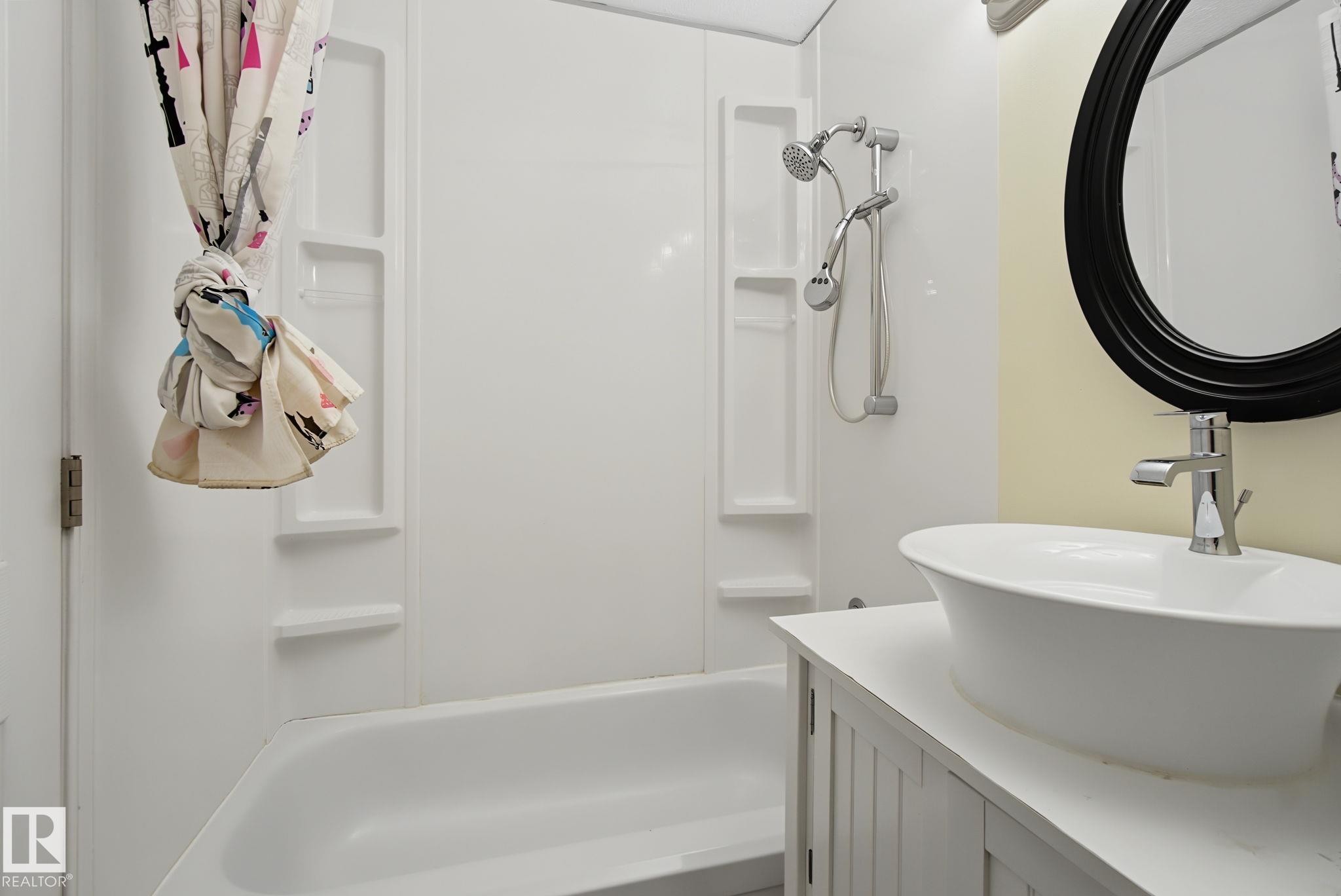 Bathroom featuring a white tub with integrated shower surround, a vessel sink with a single-handle faucet, a white vanity with cabinet storage, and an oval mirror with a dark frame - 4259 29 Avenue, Edmonton, AB