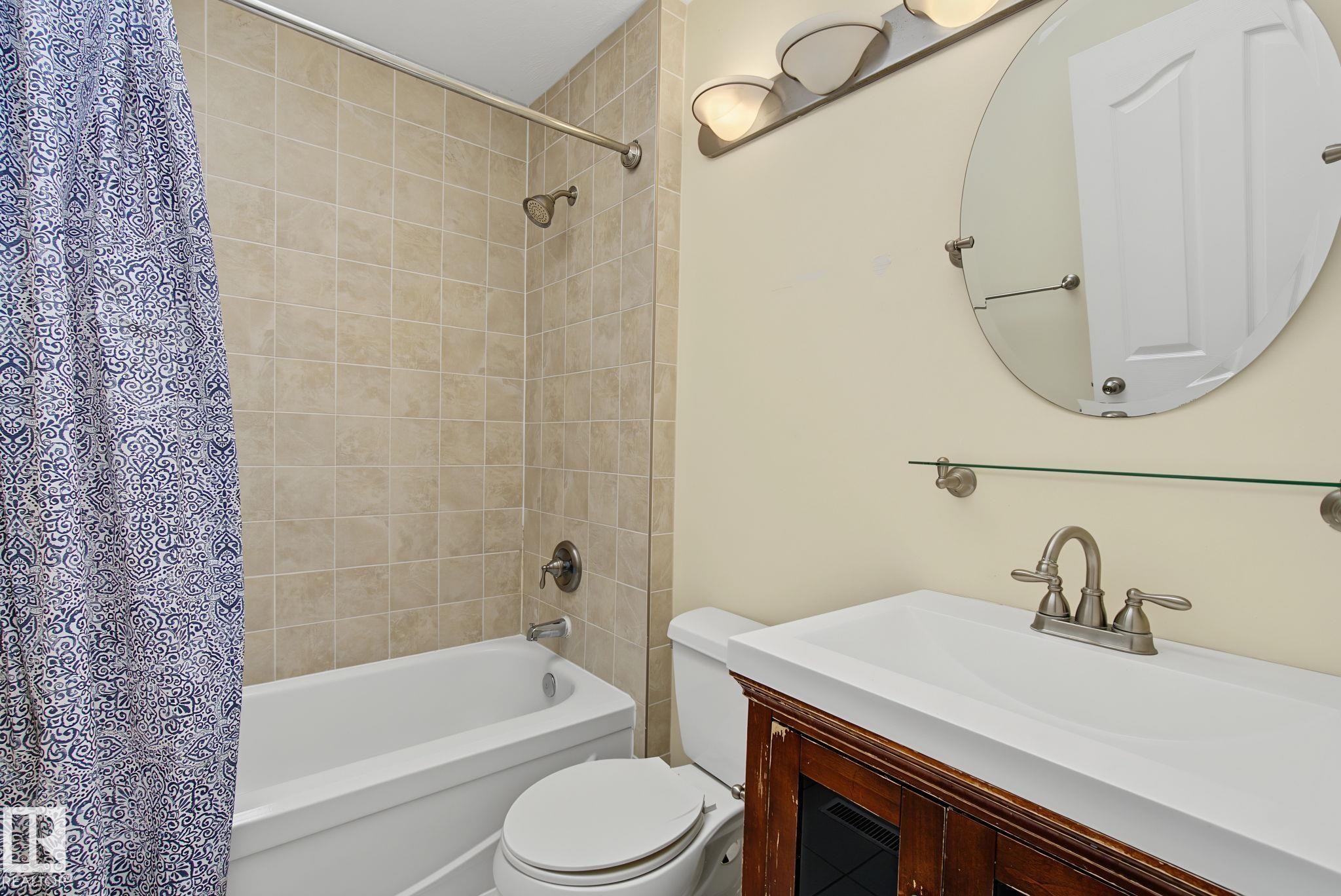 Bathroom featuring a full-size bathtub with ceramic tile surround, a built-in vanity with a white countertop, an oval wall-mounted mirror, brushed nickel fixtures, and a three-light vanity bar - 4259 29 Avenue, Edmonton, AB