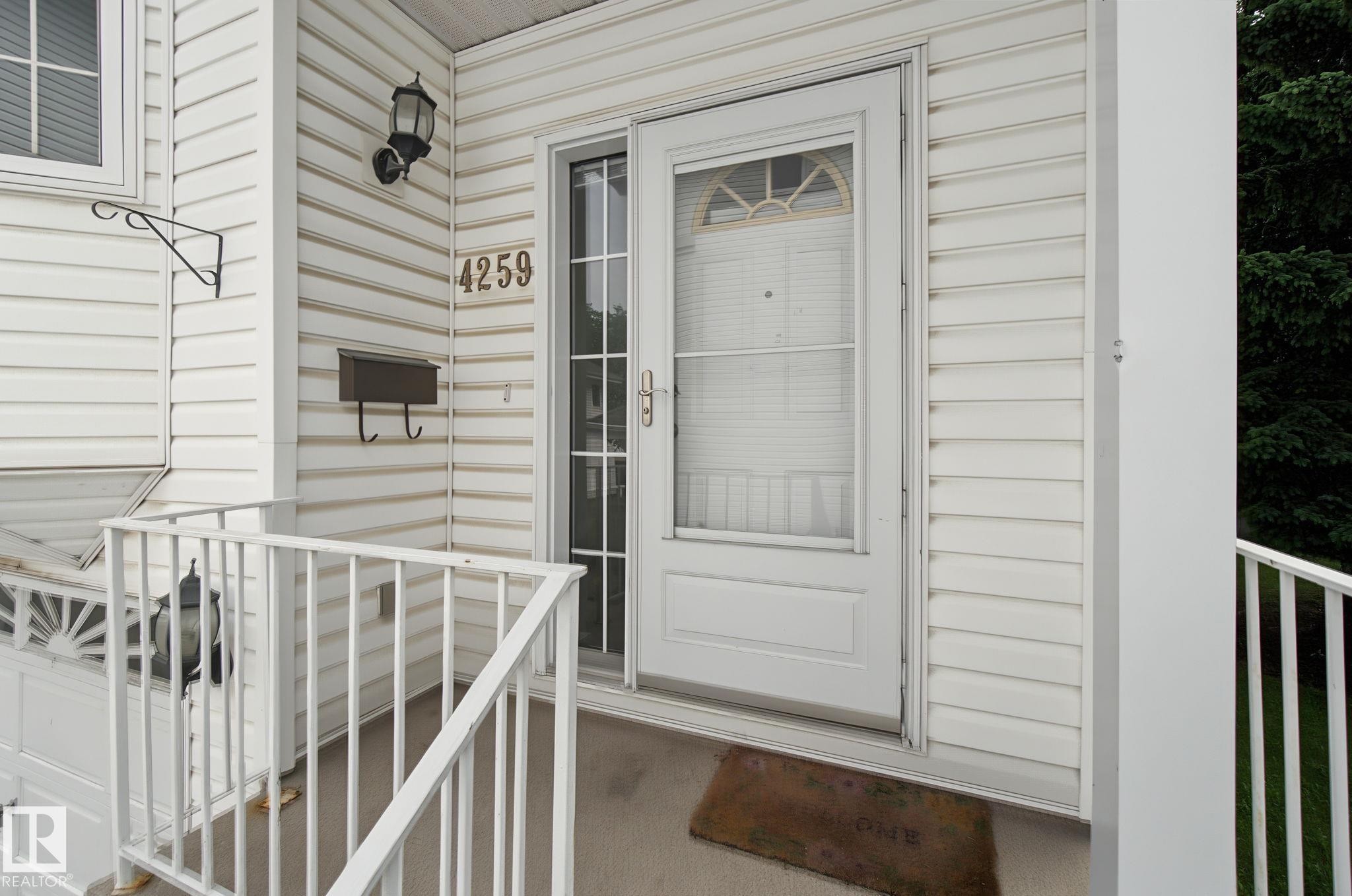 White vinyl siding exterior featuring an entryway with a storm door, an outdoor wall lantern, a wall-mounted mailbox, and white porch railings - 4259 29 Avenue, Edmonton, AB