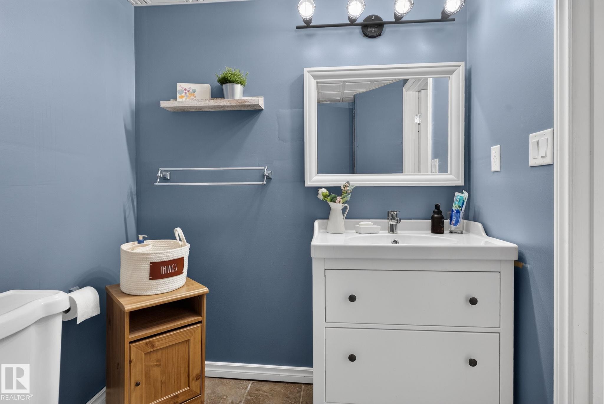 Bathroom featuring a white vanity with integrated sink, framed mirror, wall-mounted light fixture, white baseboards, and neutral flooring - 3218 41 Street, Leduc, AB