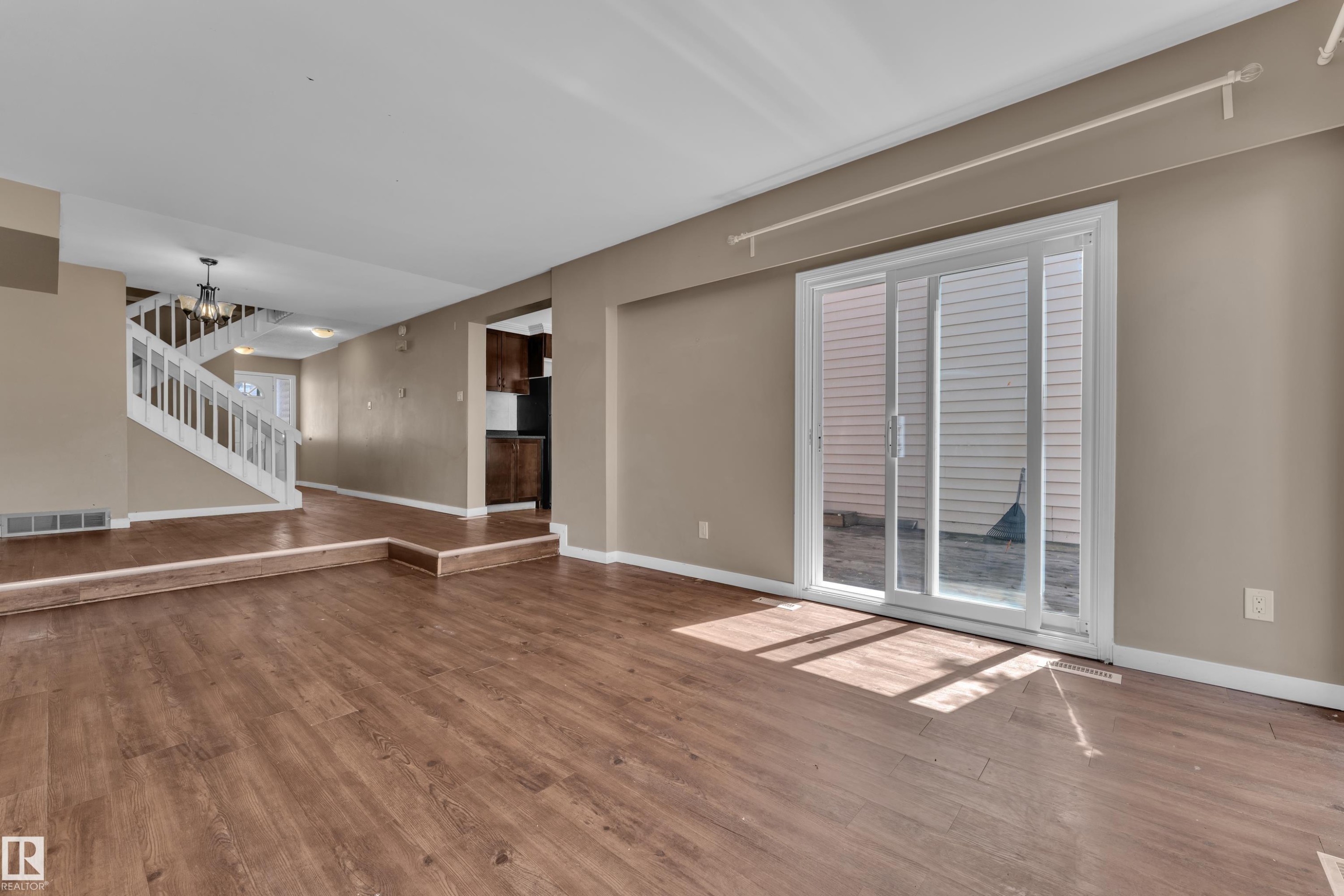 Open-concept living area with wood-finish flooring, neutral wall tones, white sliding glass door, and a multi-level floor plan leading to a staircase with white railings - 6632 36A Avenue, Edmonton, AB