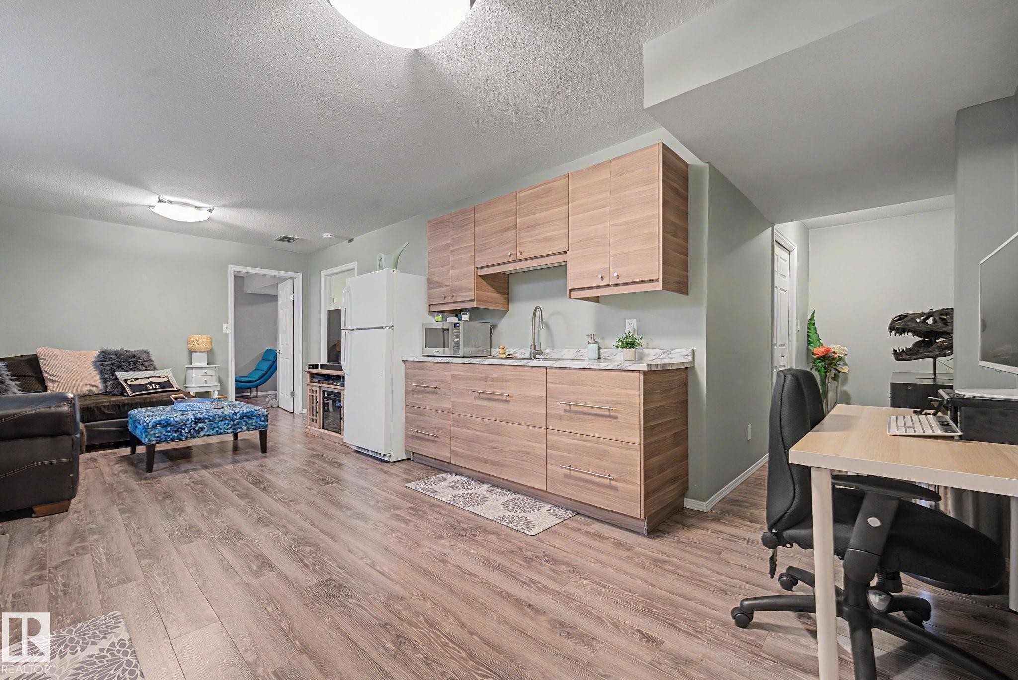 Open-concept living space featuring wood-finish flooring, a functional kitchenette with wood-tone cabinetry, a sink with a stainless steel faucet, and light-toned countertops - 19039 47 Avenue Nw, Edmonton, AB - Indoor Photo Showing Office