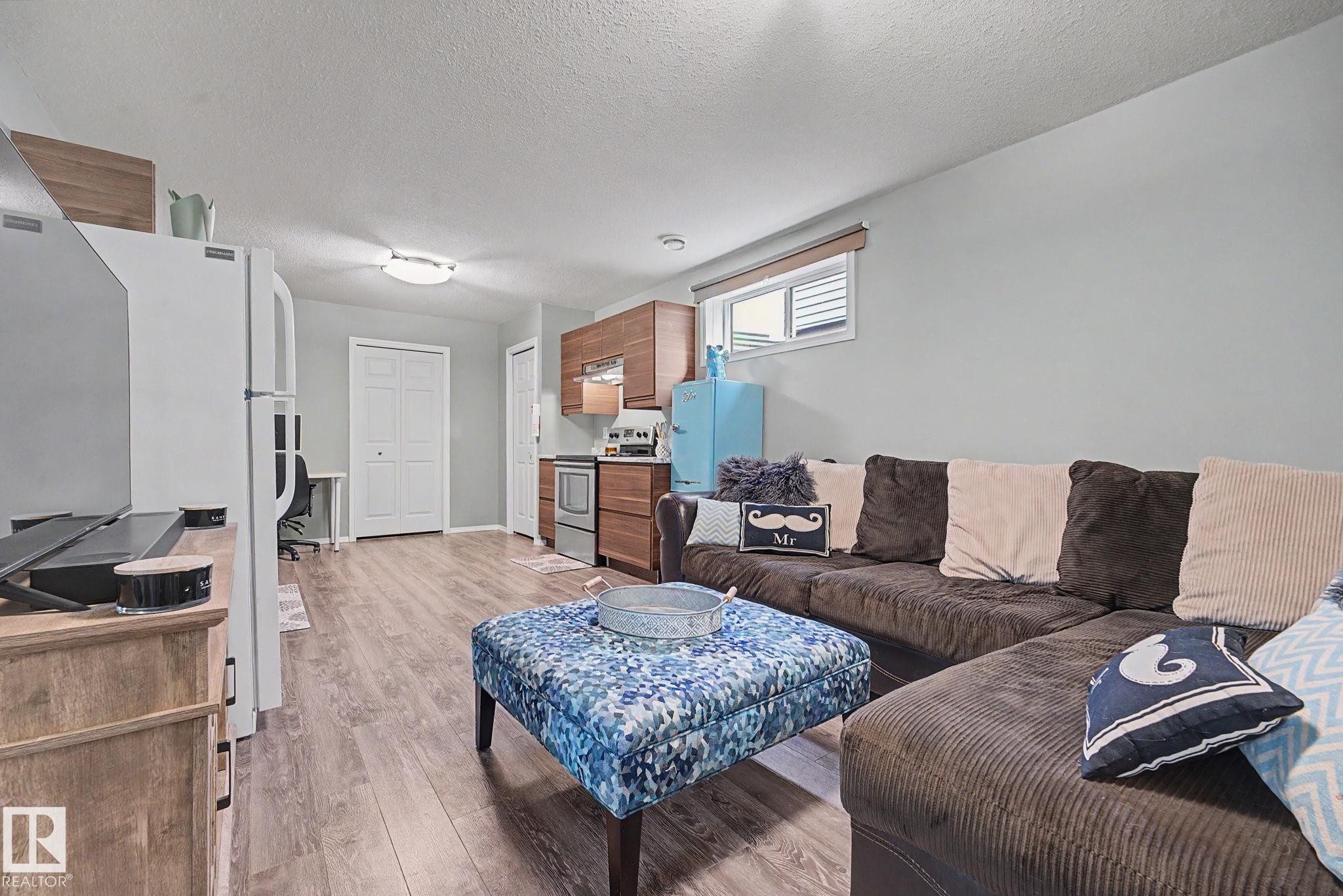 Open-concept living space featuring wood-finish flooring, a compact kitchen area with wood cabinetry, stainless steel appliances, and ample natural light from a horizontal window - 19039 47 Avenue Nw, Edmonton, AB - Indoor Photo Showing Living Room
