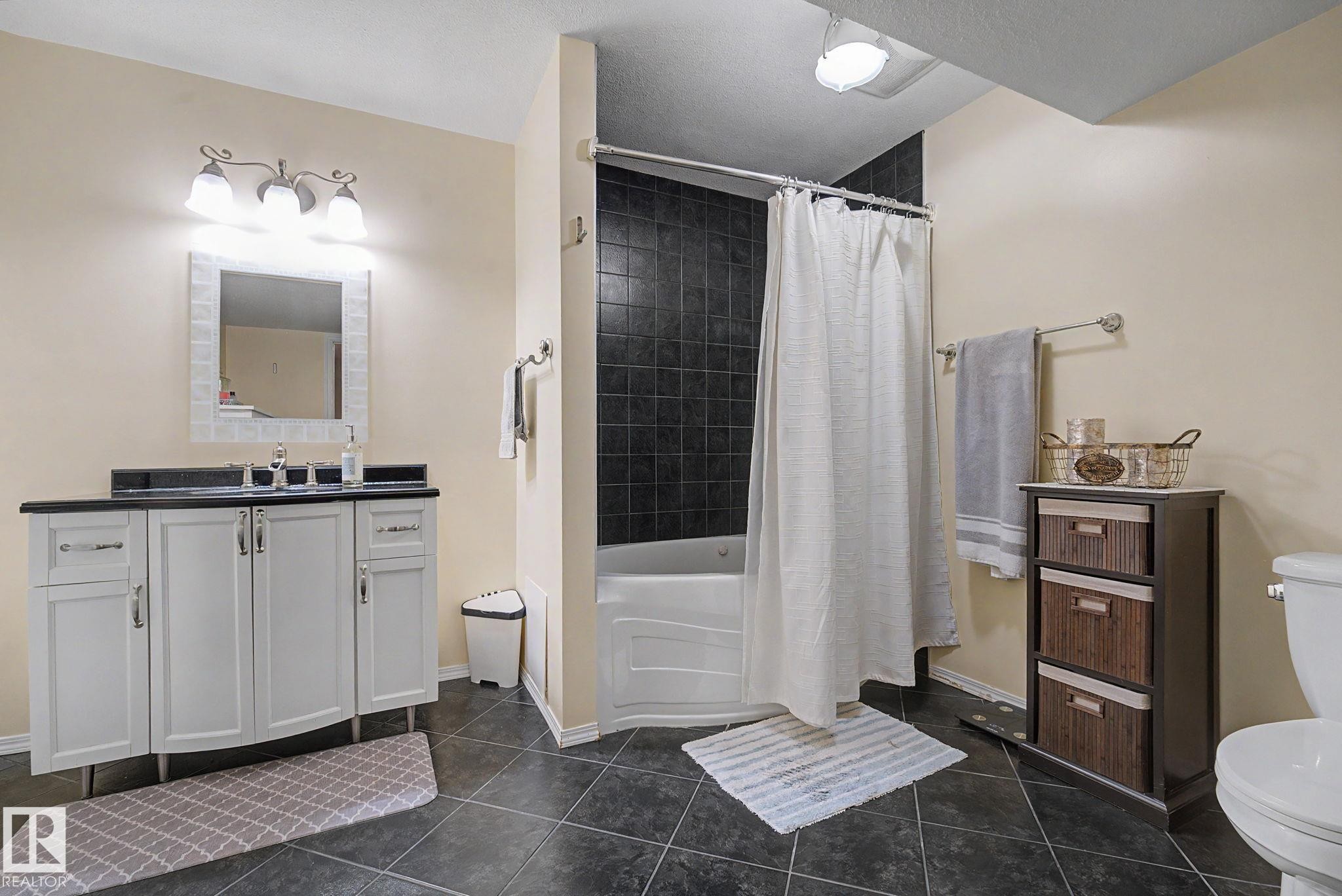Bathroom featuring dark tile flooring, a white vanity with a dark countertop, a white bathtub with dark tile surround, a wall-mounted mirror with a decorative frame, and a chrome light fixture - 19039 47 Avenue Nw, Edmonton, AB - Indoor Photo Showing Bathroom