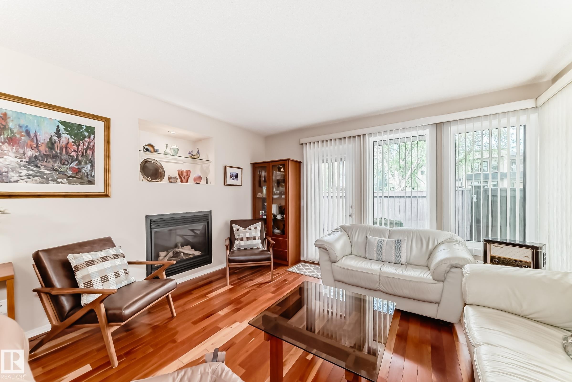 Living area featuring wood-finish flooring, a built-in fireplace, recessed shelving with glass accents, and large windows with vertical blinds - 7930 111 Street, Edmonton, AB - Indoor Photo Showing Living Room With Fireplace