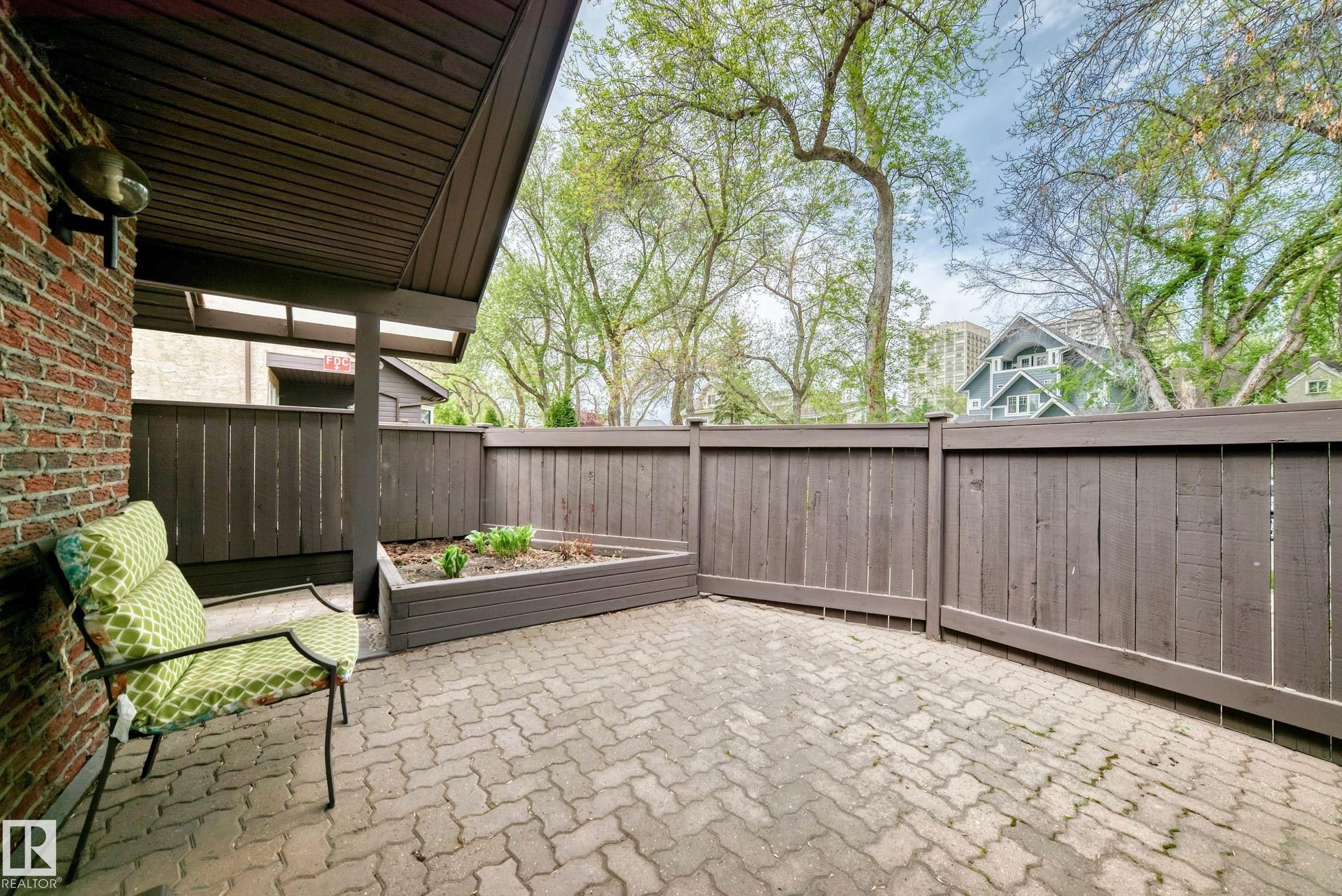 Private patio, accessed from living room. Paved patio featuring a raised garden bed, brown privacy fencing, and a brick exterior wall with an attached outdoor light fixture - 7930 111 Street, Edmonton, AB - Outdoor With Deck Patio Veranda With Exterior