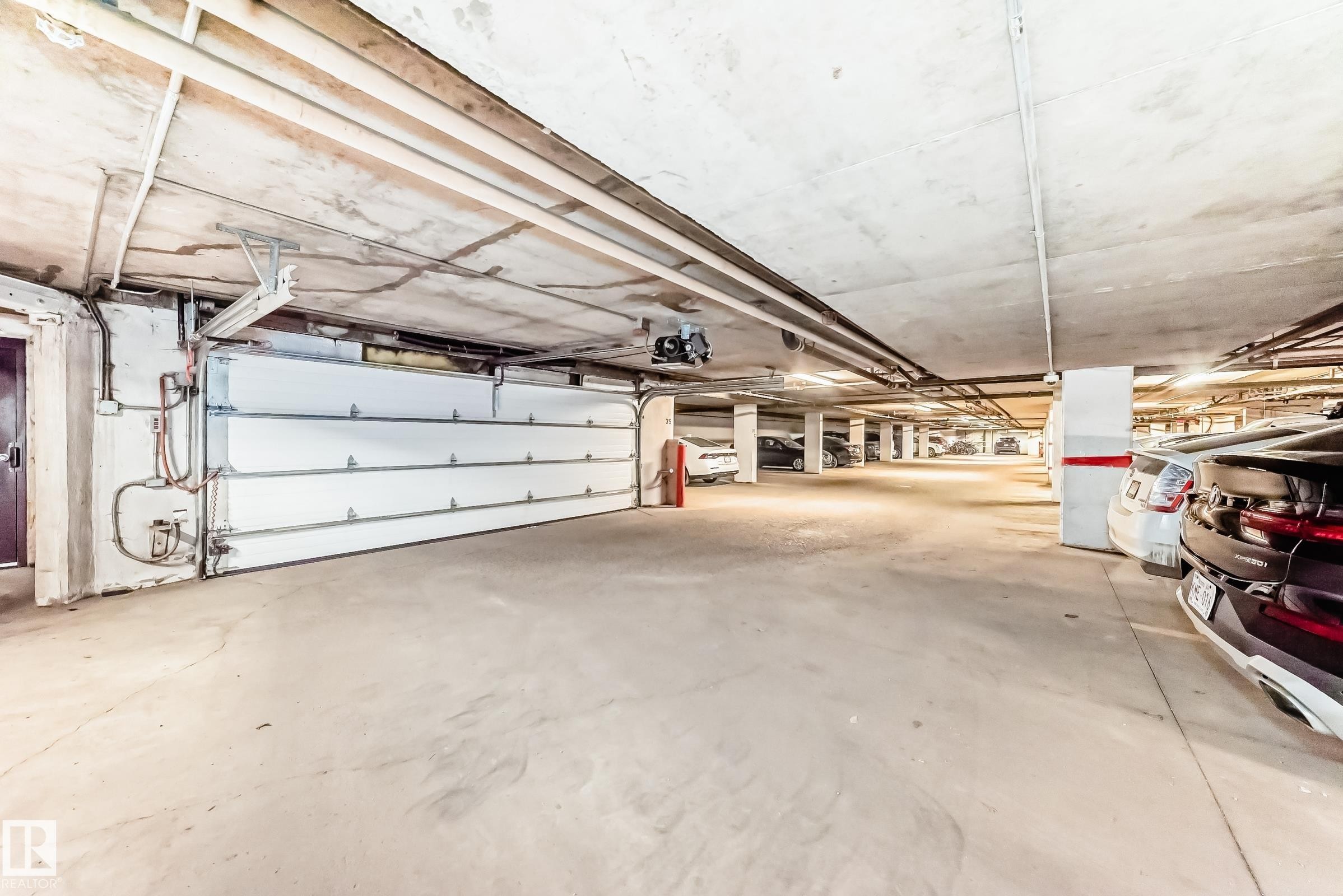 Underground parking garage featuring a white sectional garage door, concrete flooring, and exposed ceiling utilities - 7930 111 Street, Edmonton, AB - Indoor Photo Showing Garage