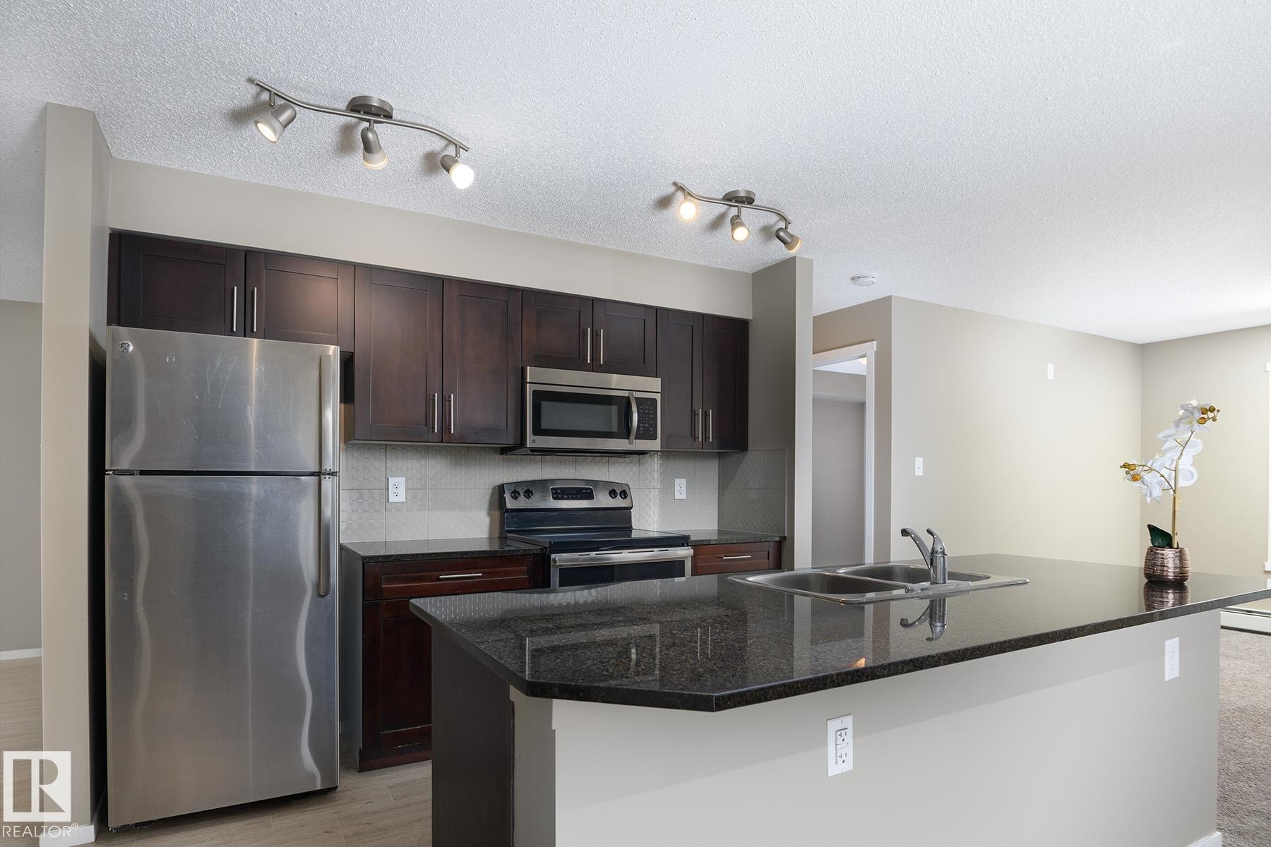 Kitchen featuring dark wood cabinetry, stainless steel appliances, a tile backsplash, and a breakfast bar with a double basin sink - 102 25 Element Dr, St. Albert, AB