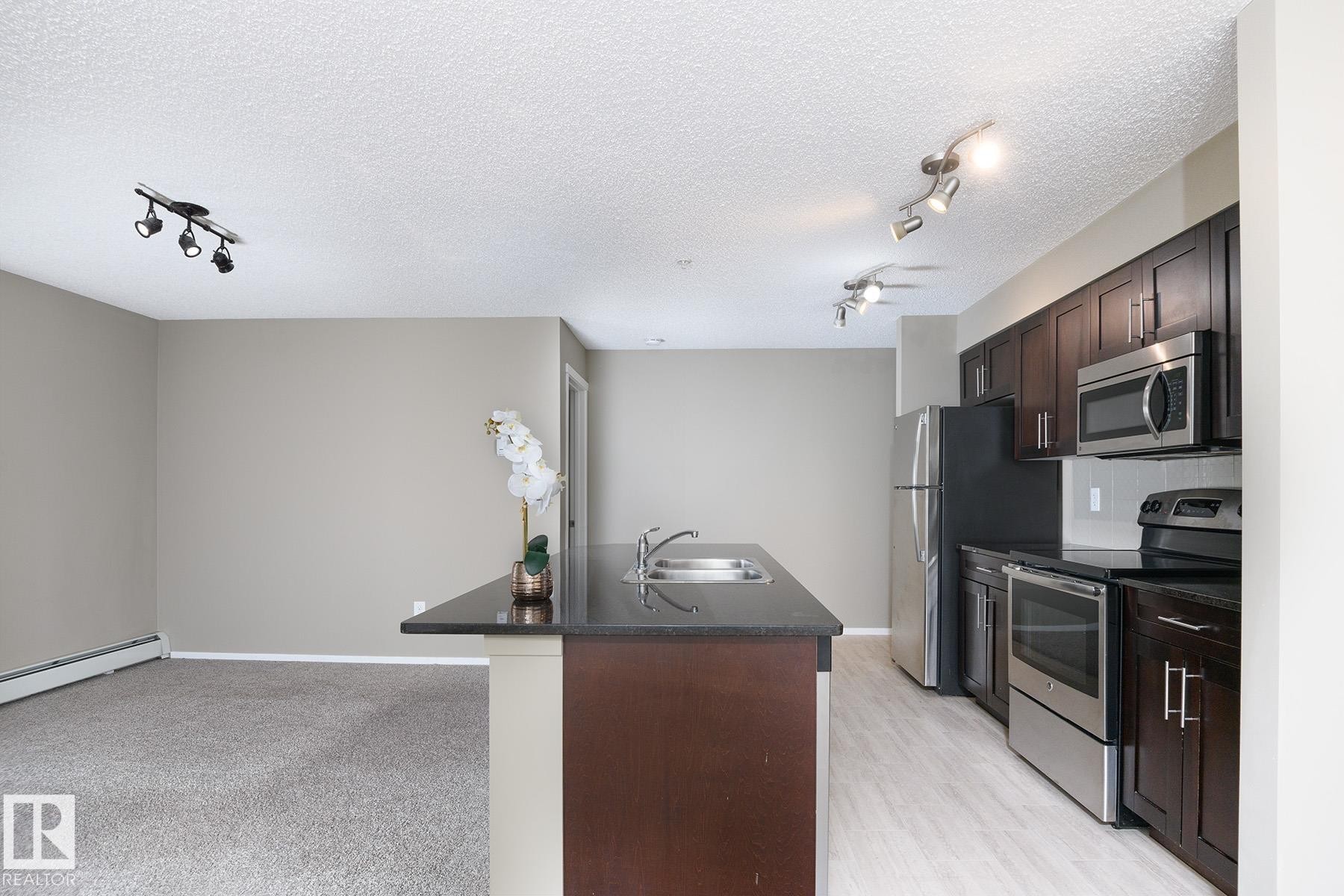 Kitchen featuring dark wood-finish cabinetry, stainless steel appliances, and a central island with an integrated sink and dark countertop - 102 25 Element Dr, St. Albert, AB